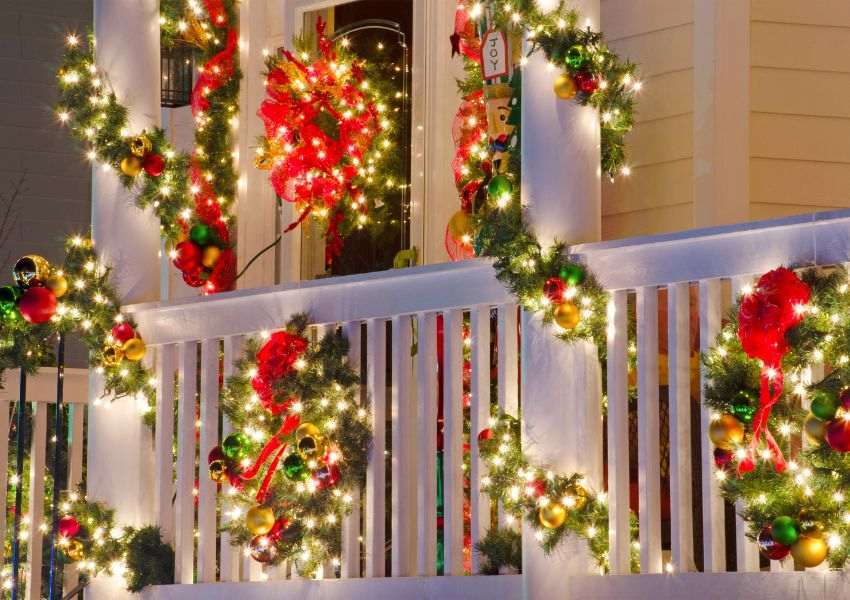 A close up of a pale yellow HOA property with white banisters is decorated with green and red holiday decorations with warm yellow lights and colorful bells throughout.