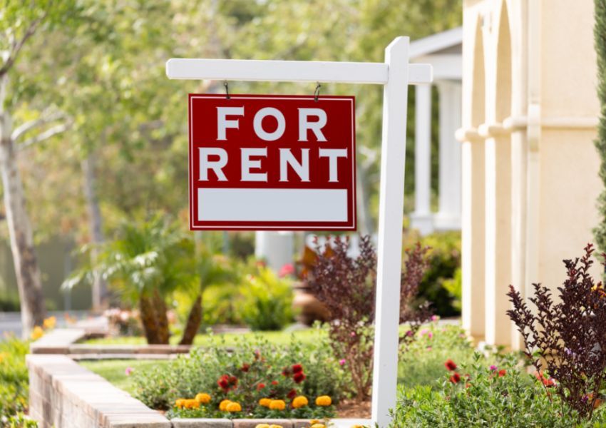 A red and white ‘for rent’ sign hangs from a white wooden post in front of a rental property’s garden.