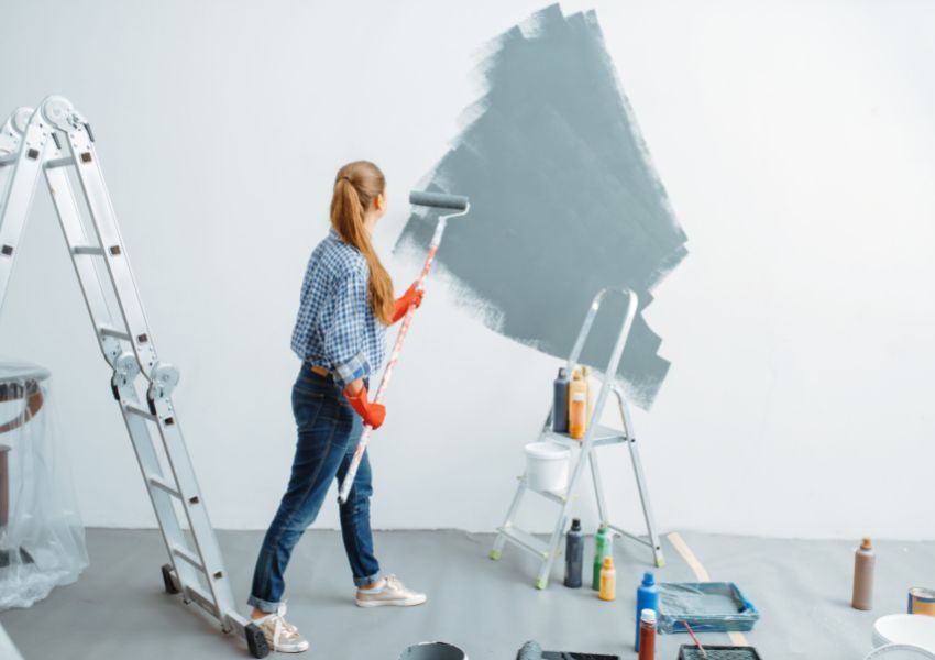 A landlord with a long brown ponytail and plaid shirt paints a rental unit’s wall grey with a painting roller; a ladder, stepladder and painting supplies surround them.