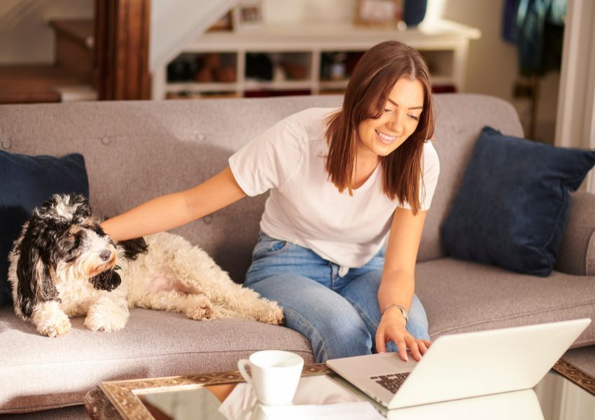 A smiling HOA community member with long brown hair sits on a grey couch in their home with a black and white dog beside them as they look at their laptop.