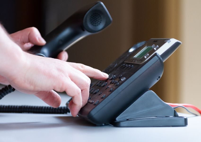 A landlord’s right hand dials a property manager’s number into a black landline on a desk while holding the phone in their left hand.
