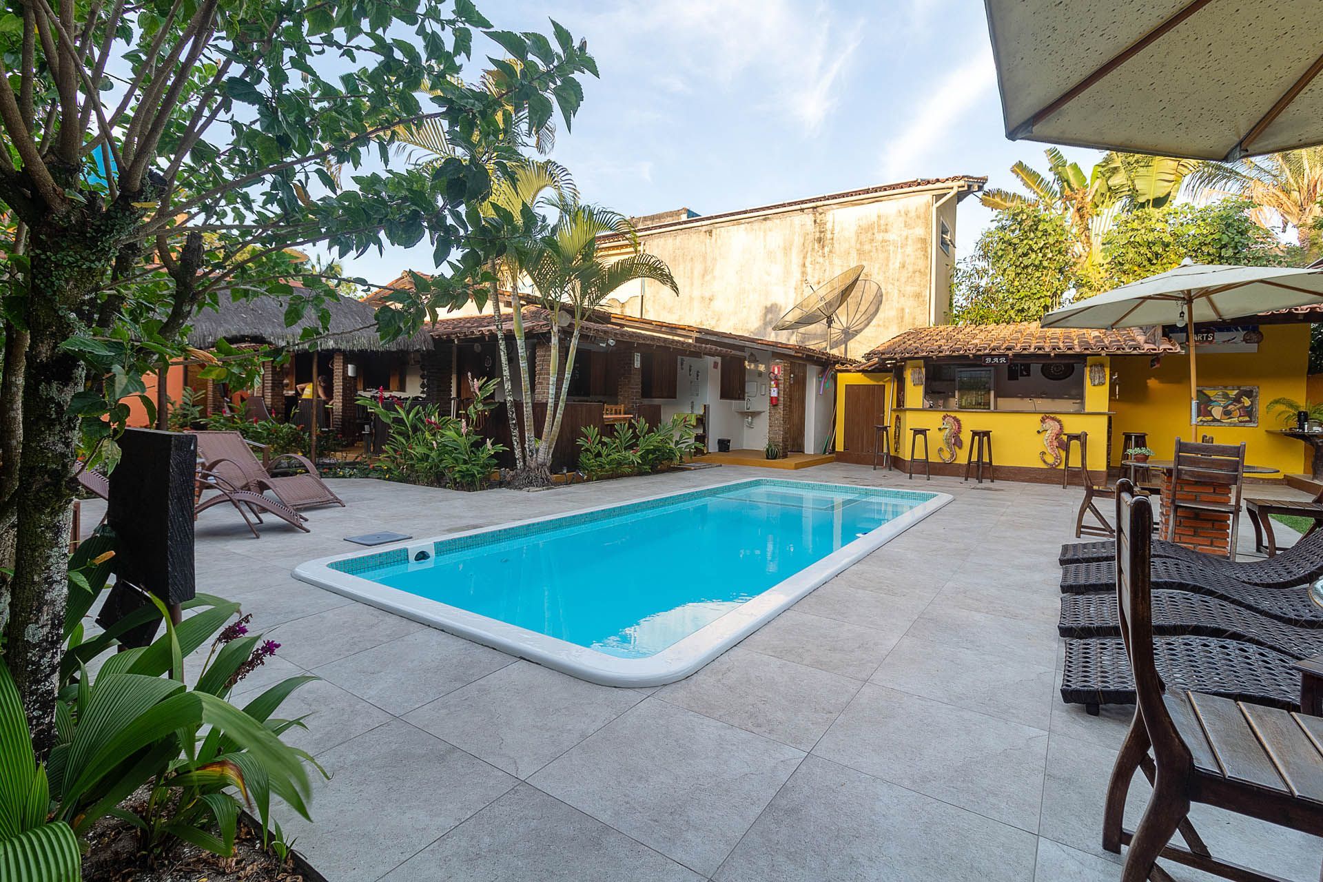 Piscina no pátio com água azul, terraço com azulejos, espreguiçadeiras e árvores tropicais ao lado de edifícios amarelos.
