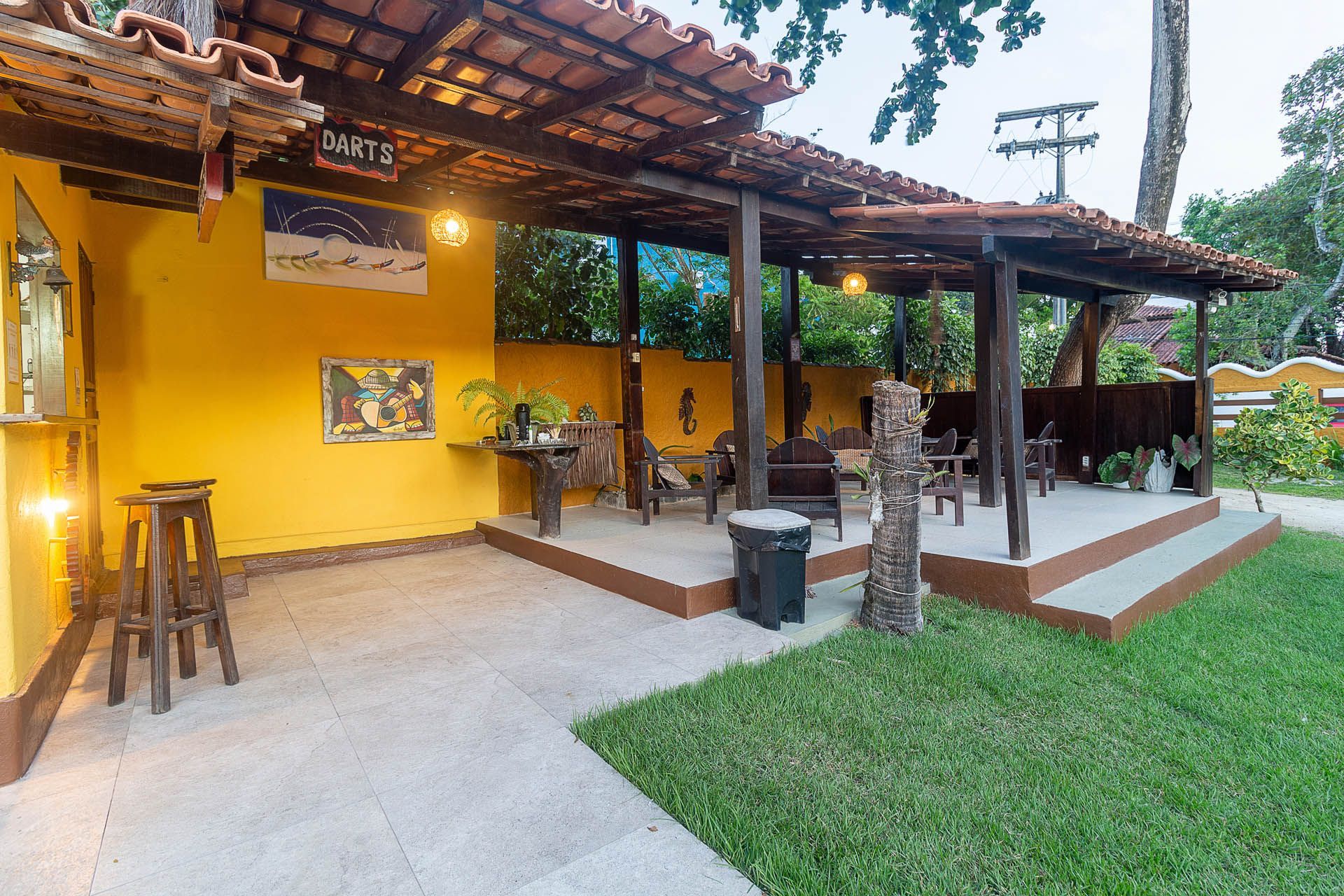 Pátio externo amarelo com pérgola de madeira, área de estar e gramado verde em um ambiente tropical.
