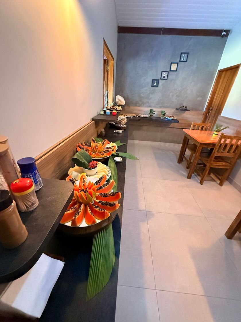 Mesa de buffet com travessas de frutas e pratos em uma sala de jantar iluminada com cadeiras de madeira.