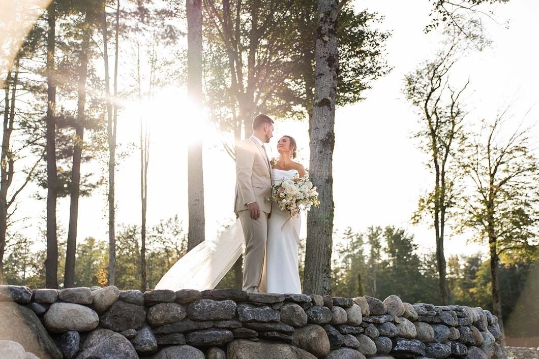A bride and groom are standing on a stone wall in the woods.