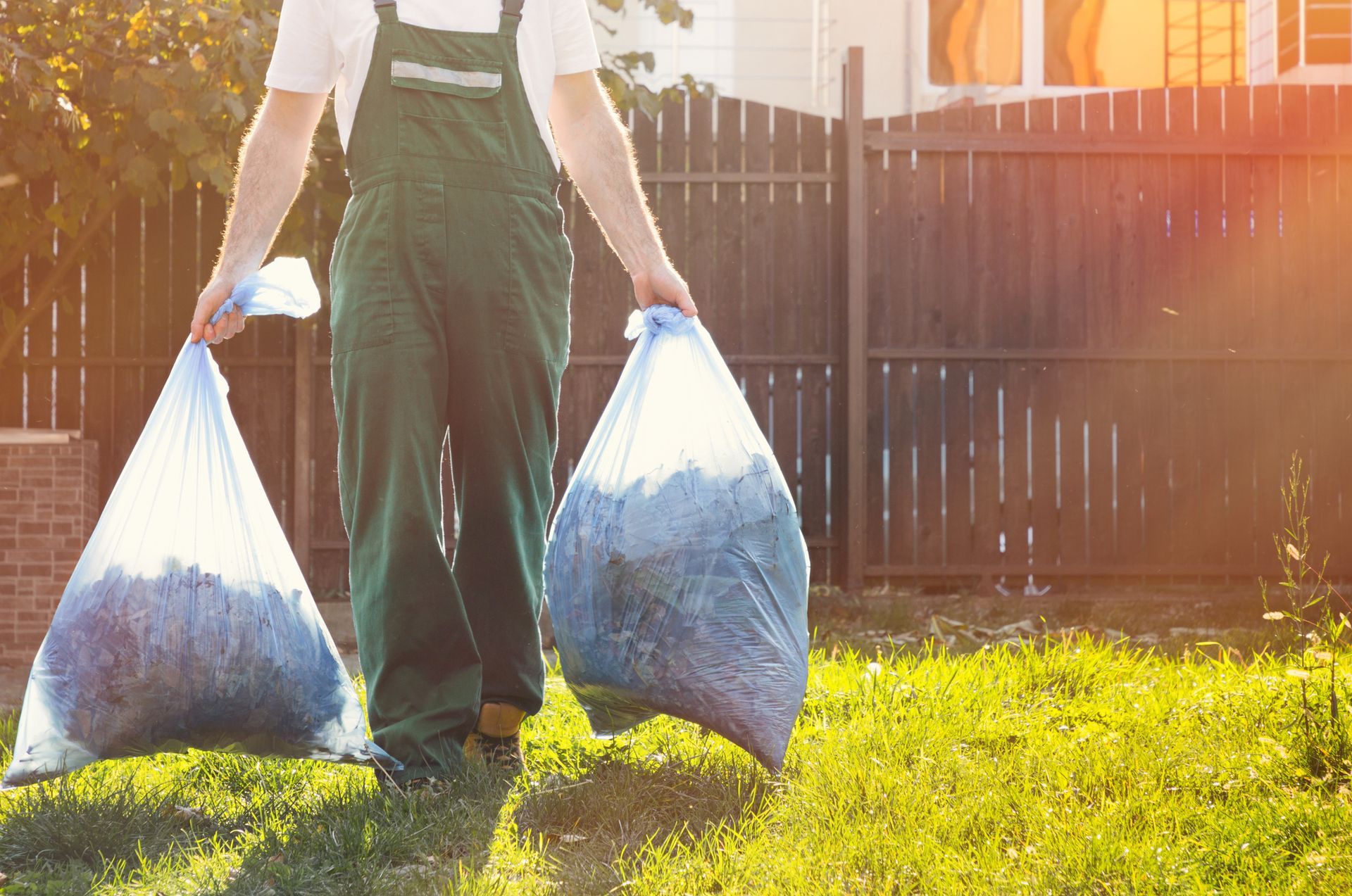 A man is carrying two bags of trash in his backyard.