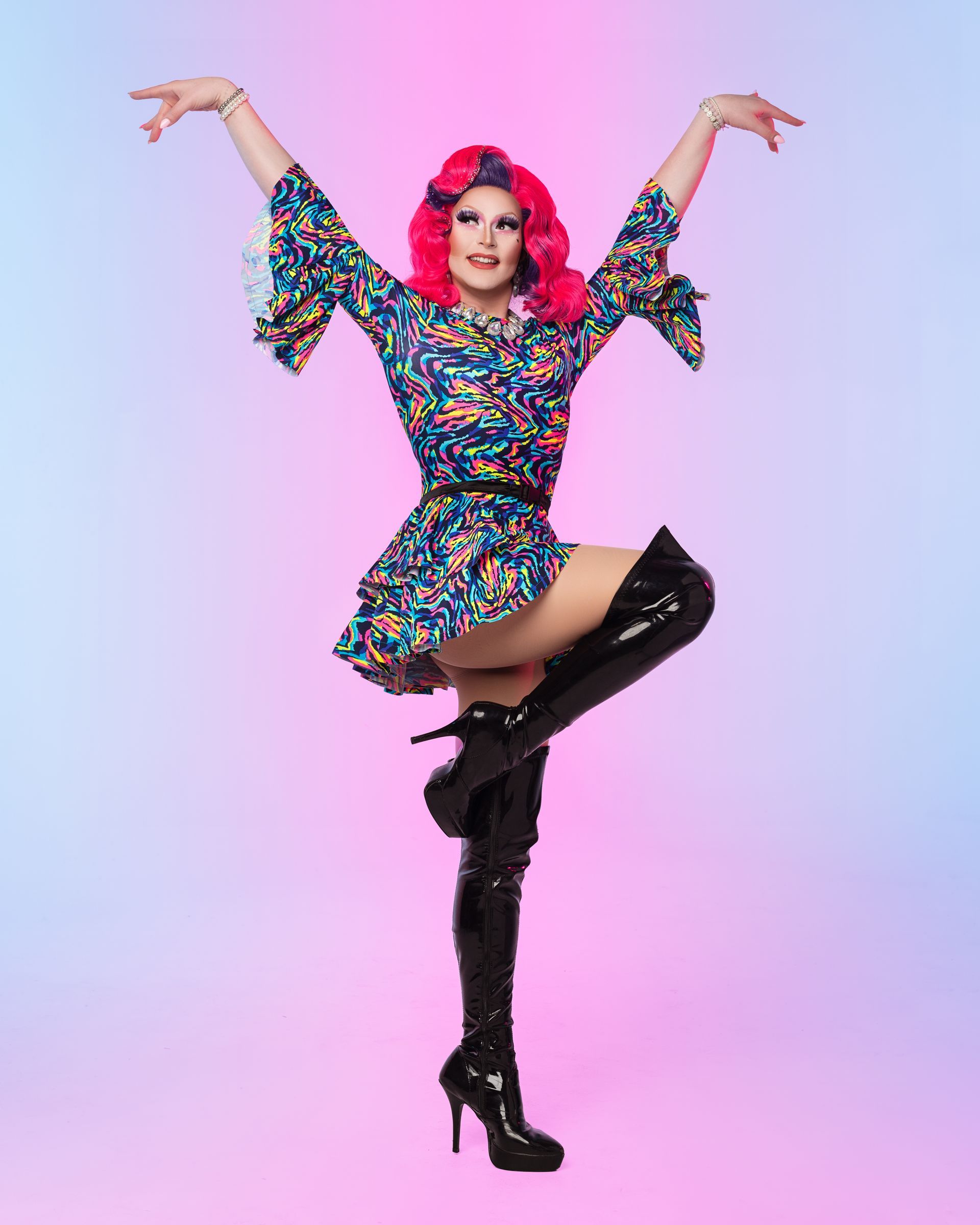 Drag queen in colorful dress and thigh-high boots, striking a pose with arms up and leg extended. Pink wig.