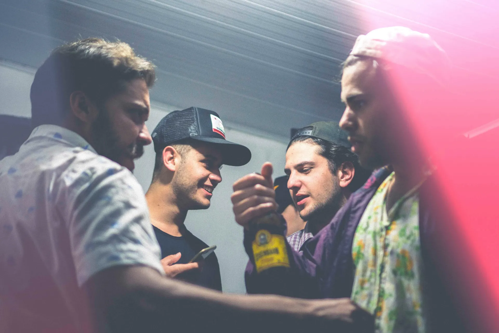 Four men in a dimly lit setting, one holding a bottle, talking and smiling.