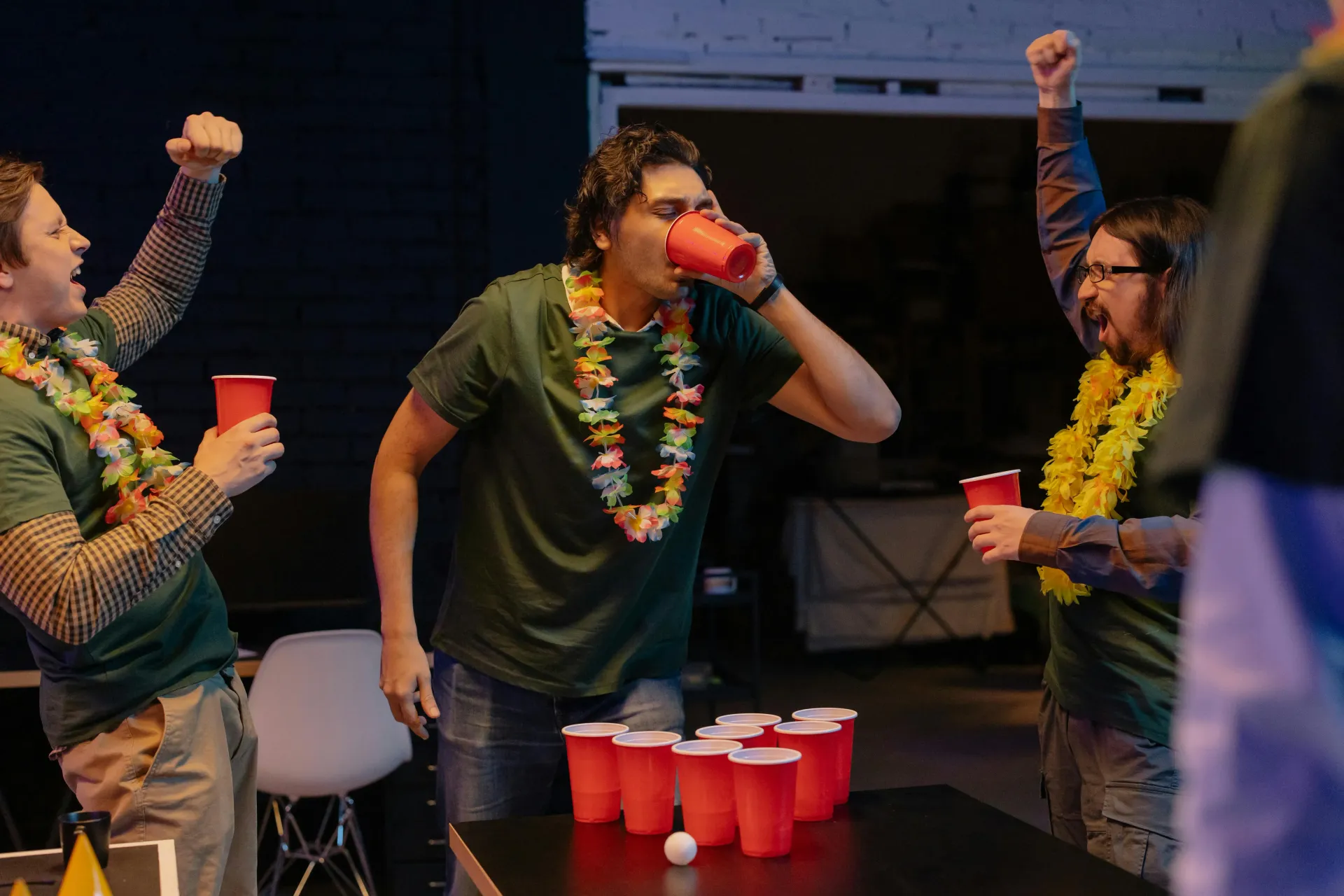 Three people wearing leis celebrate while playing beer pong, with one drinking from a red plastic cup.