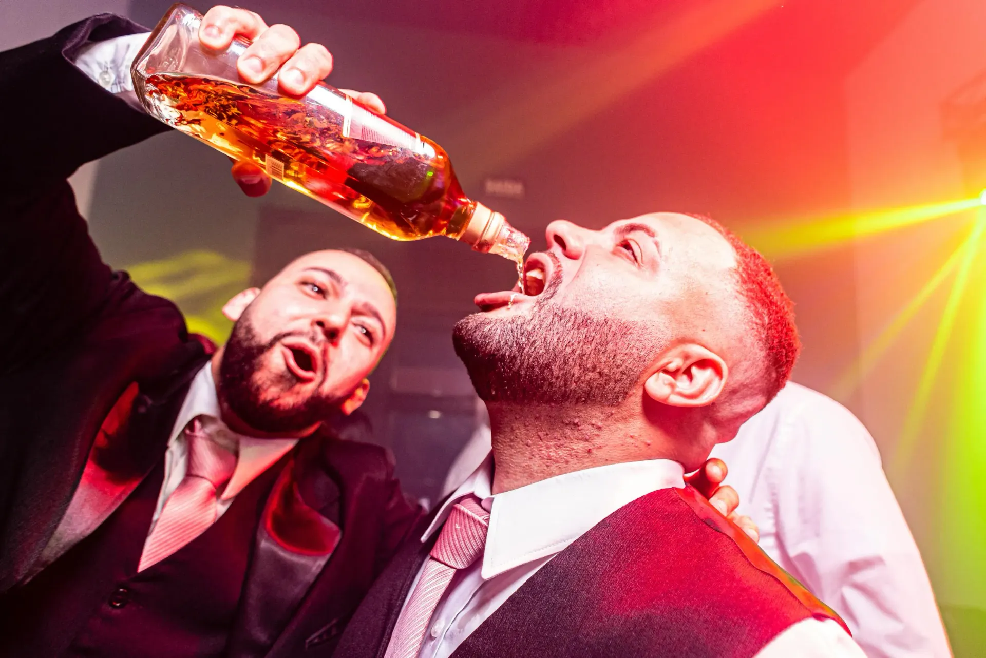 Two men drinking from a liquor bottle at a bucks party, bright lights in background.
