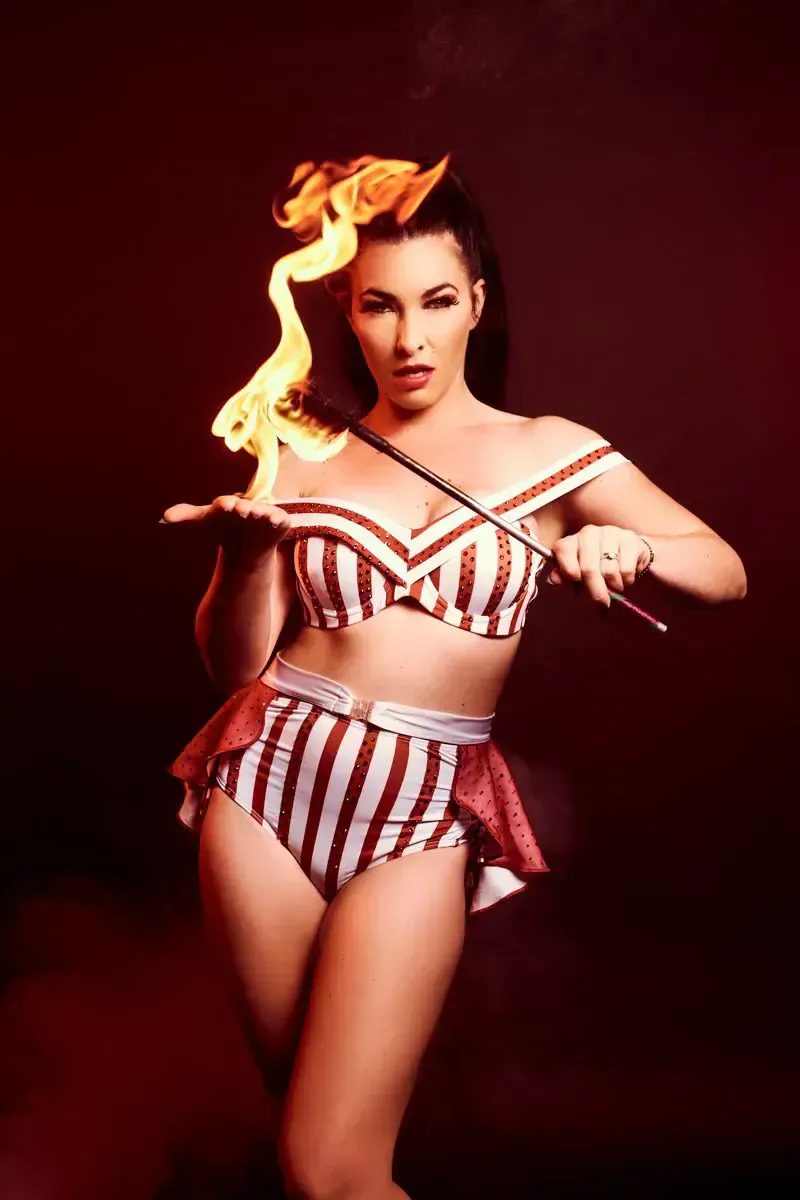 Fire dancer wearing red stripped costume, holding stick on fire, looking at the camera.