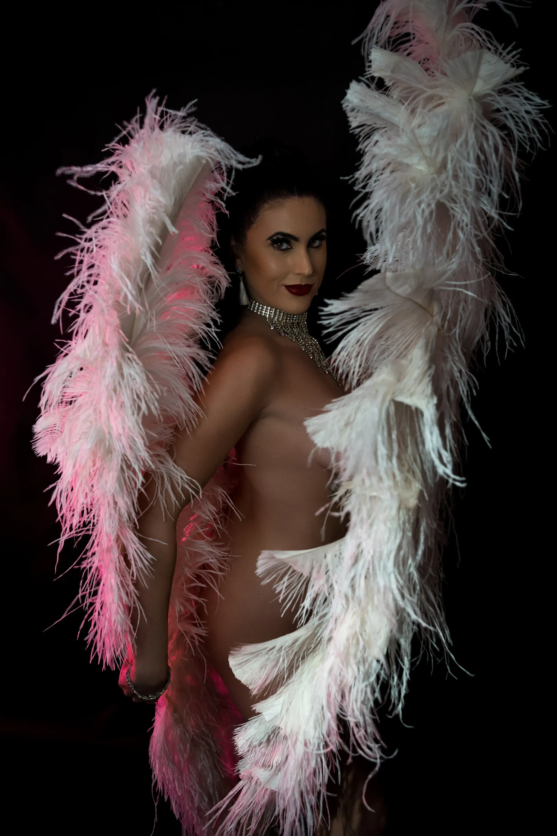 Glamour burlesque performer posing with large white feather fans and sparkling choker against dark backdrop.