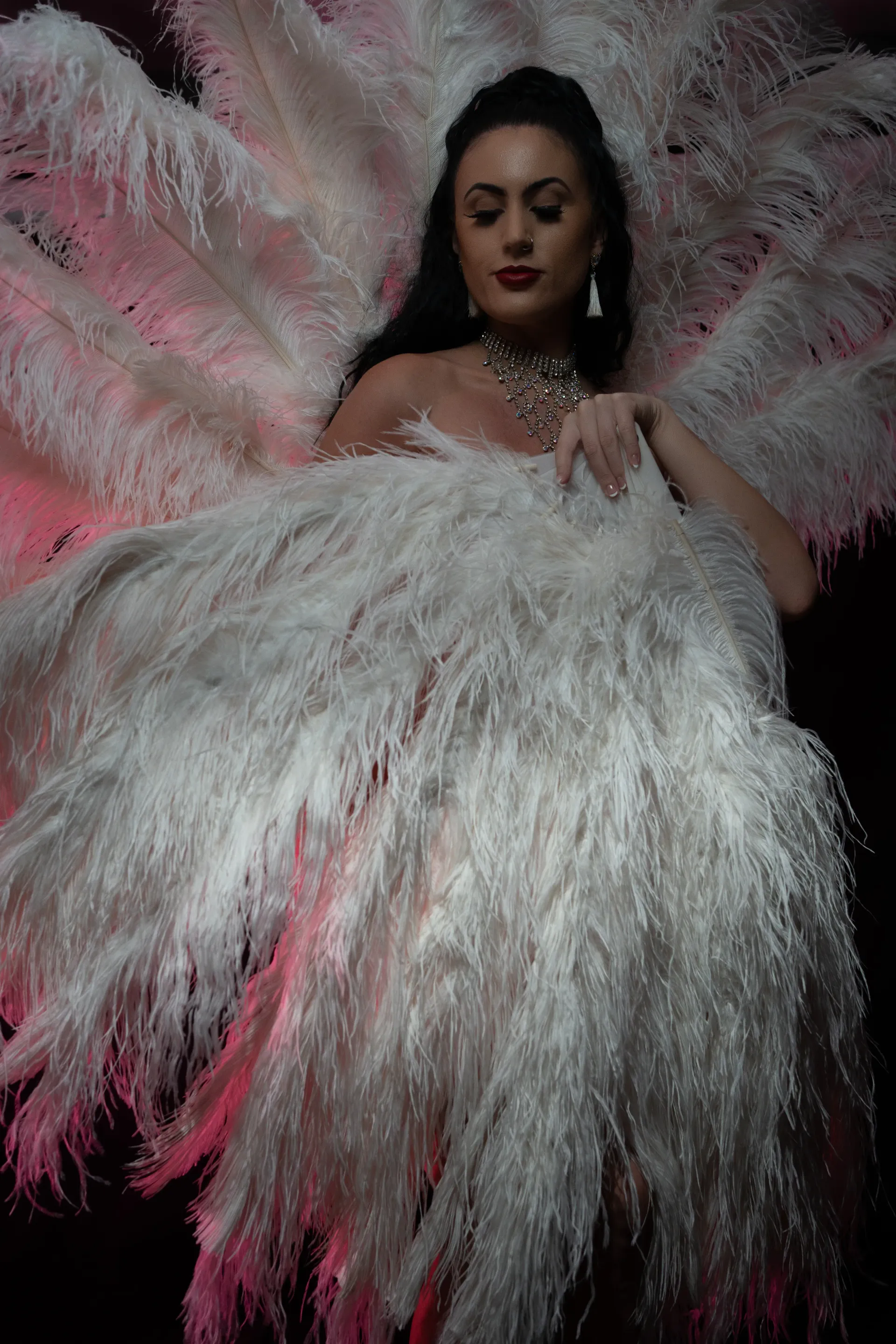 Burlesque performer covering herself with white feather fans in a pink studio lighting.