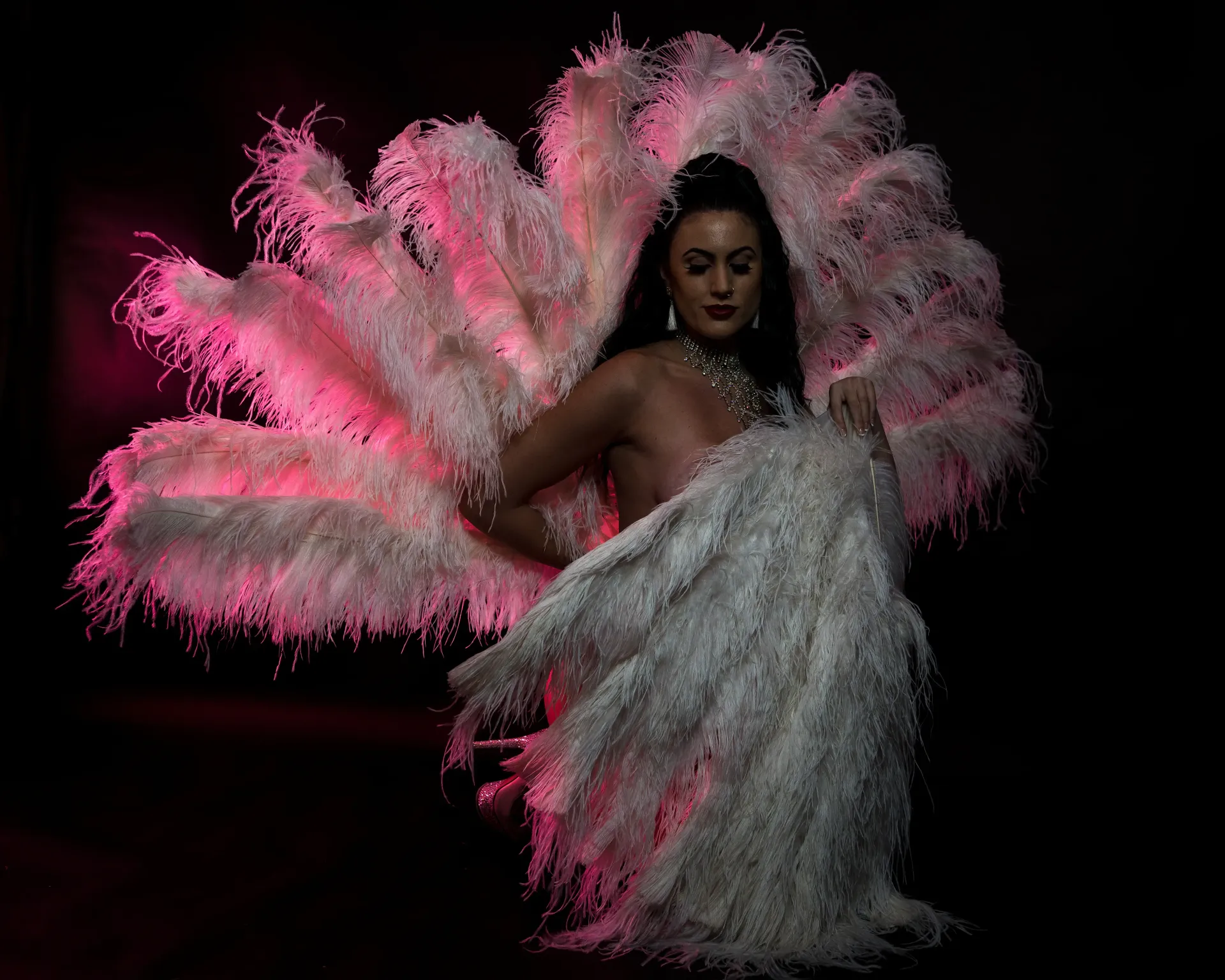 Burlesque performer in white feathery costume with pink lighting; dark background.