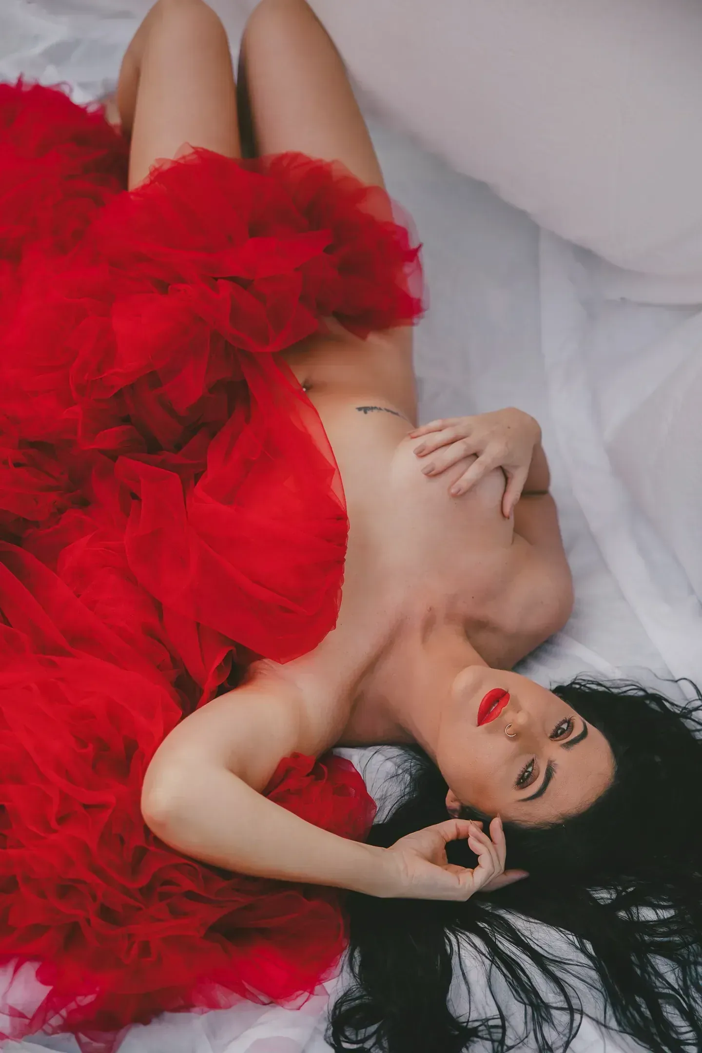 Burlesque performer reclining on white bedding, draped in dramatic red tulle with bold red lipstick for a classic pin-up look.