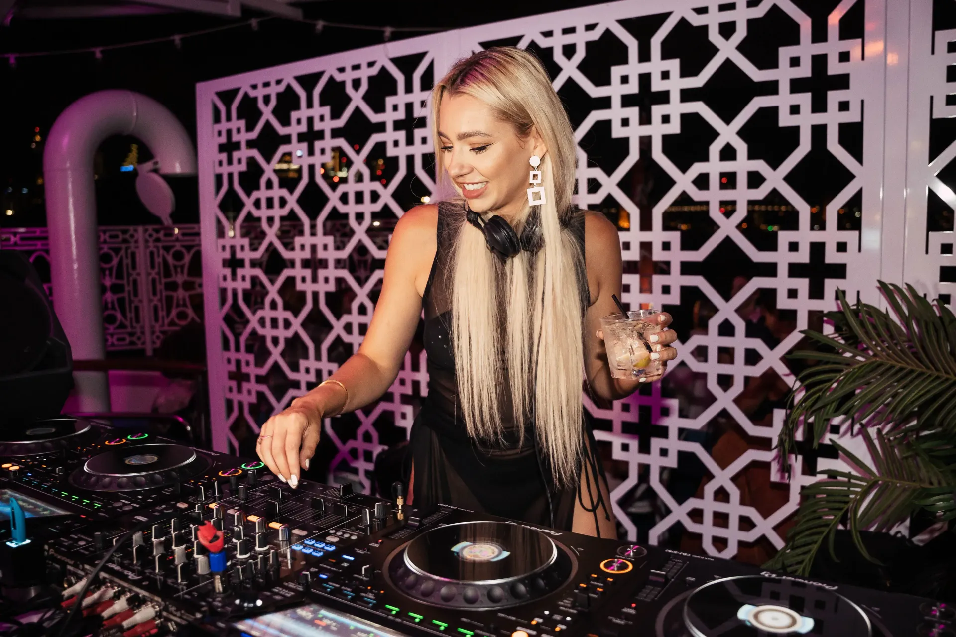 DJ at a party, blonde hair, dark outfit, smiling, mixing music on turntables, outdoor setting.