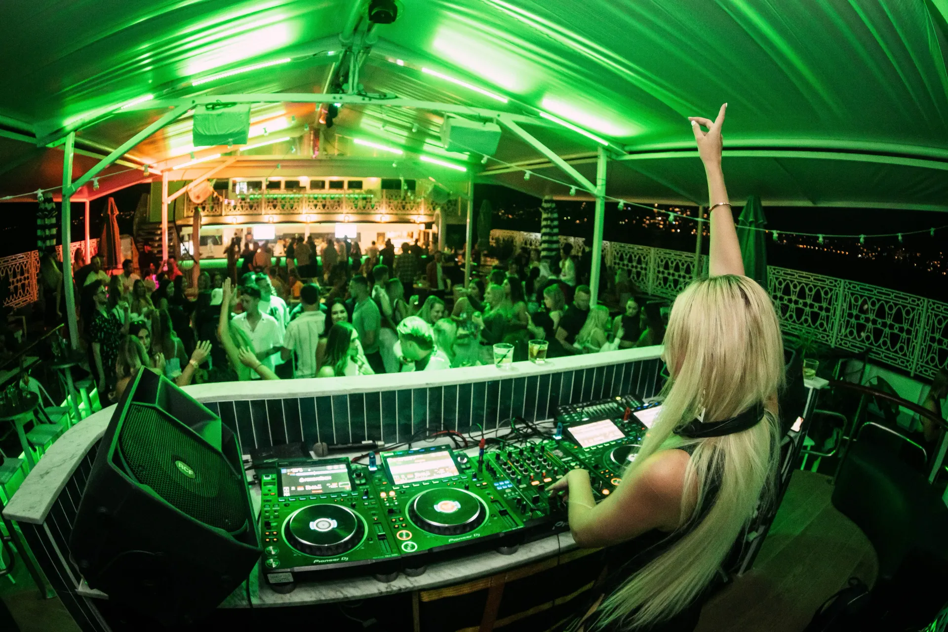 Blonde DJ with mixing on DJ set at green-lit outdoor club, crowd dancing.
