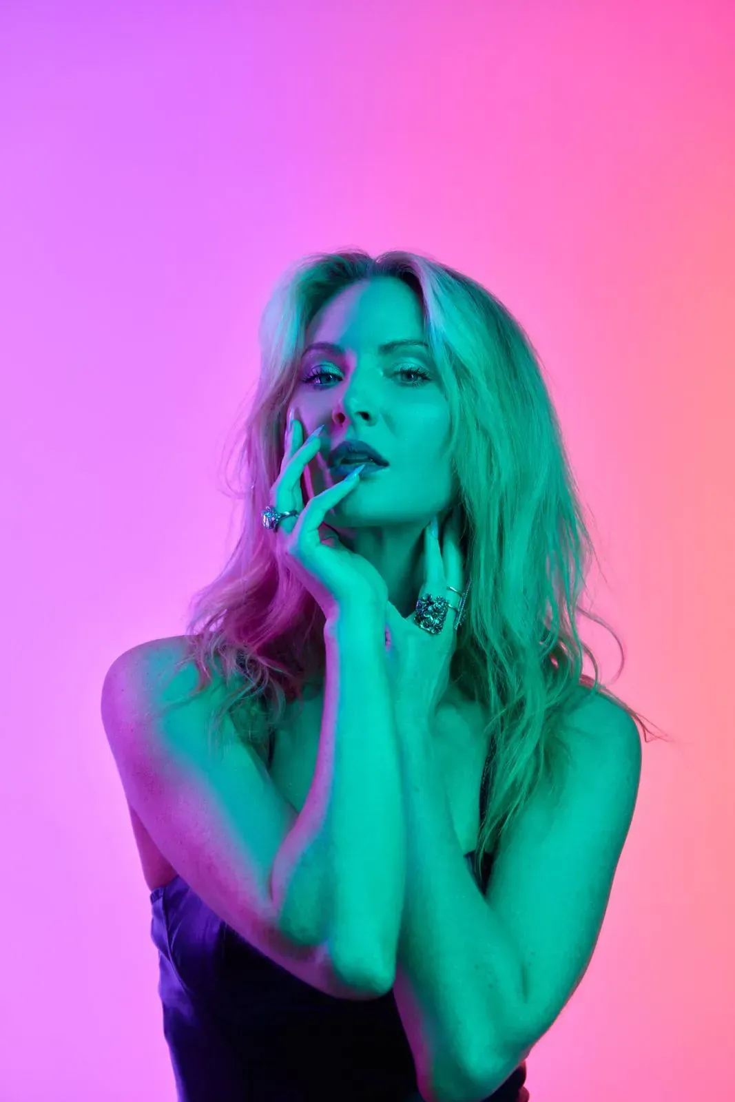 Blonde woman posing infront of gradient pink background, wearing a satin outfit and statement rings.