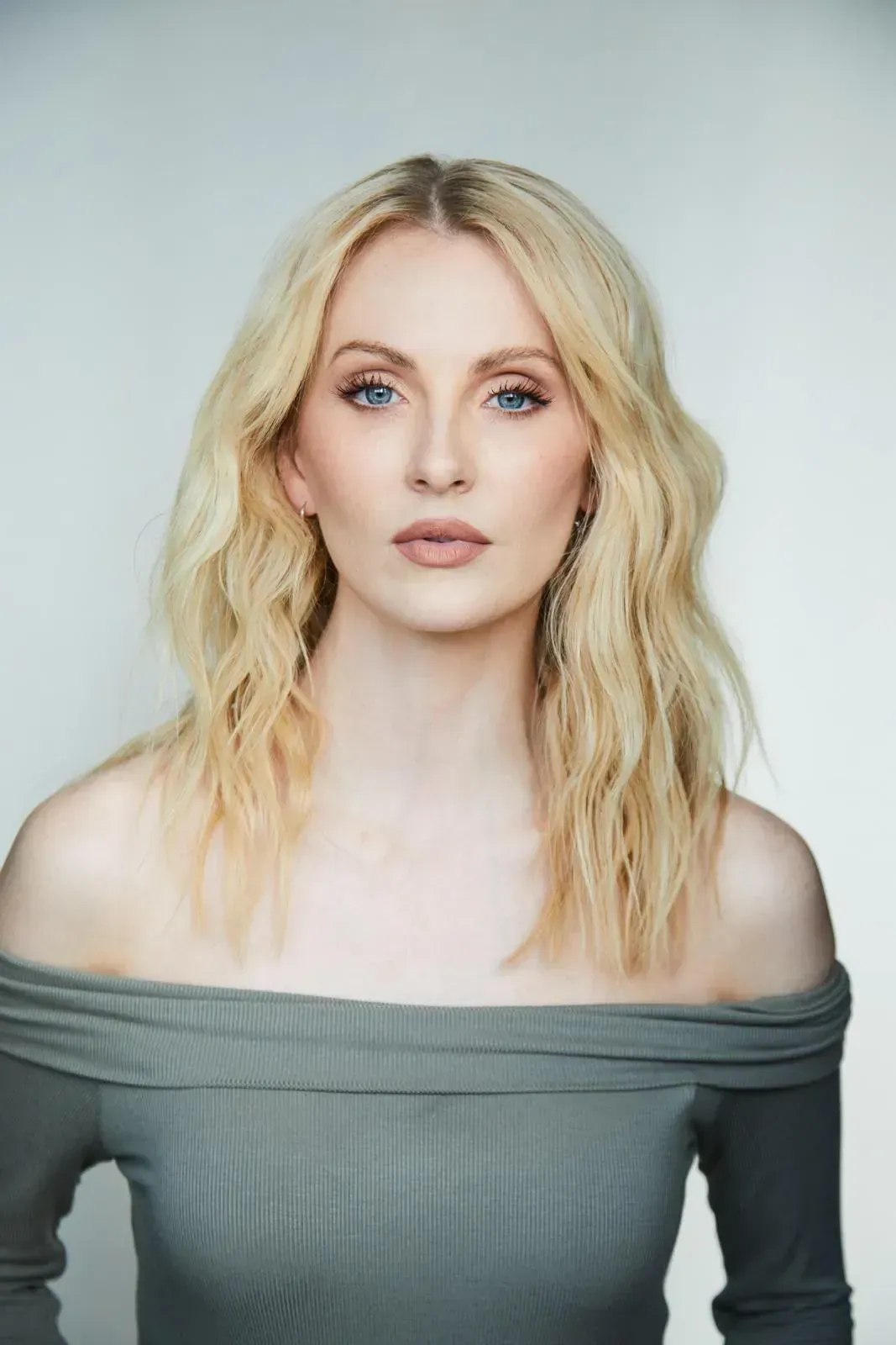 Blonde woman with wavy hair wearing an off-the-shoulder grey top, posing against a light grey background.
