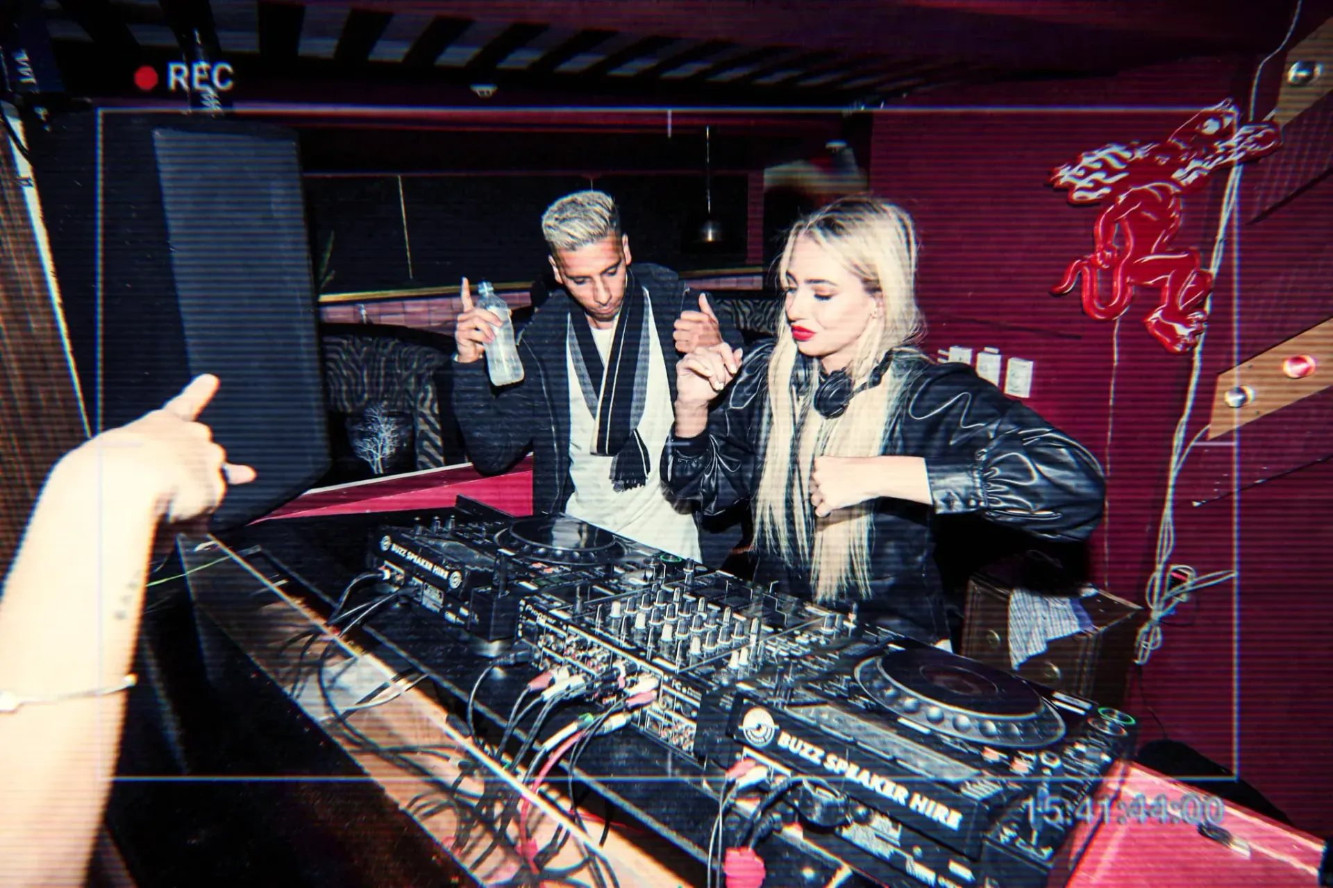 Two DJs at a club: blonde woman, man with bleached hair. Mixing music at console; hand holding drink.