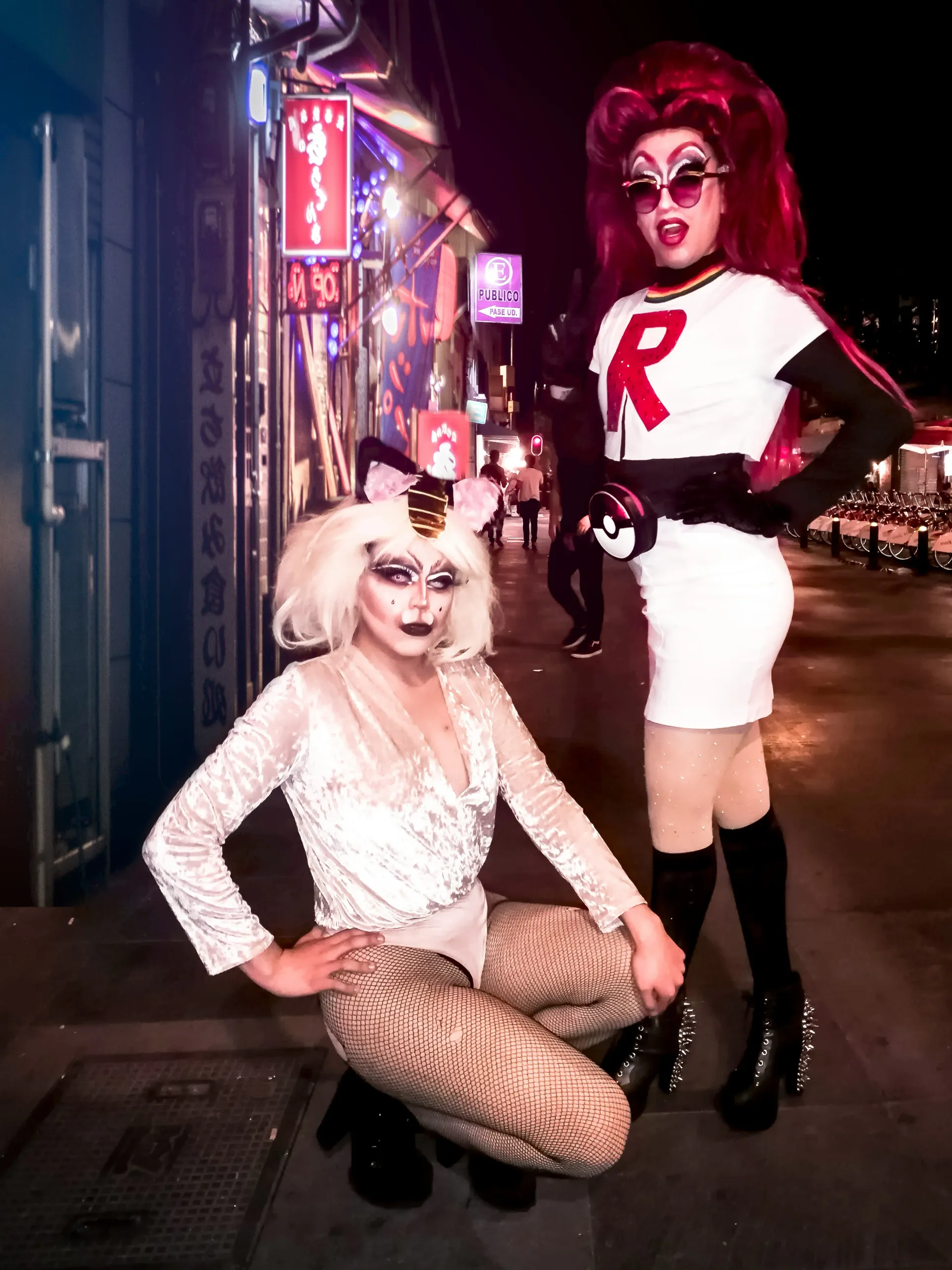 Two drag performers in bold makeup and costumes posing on a neon-lit city street at night.