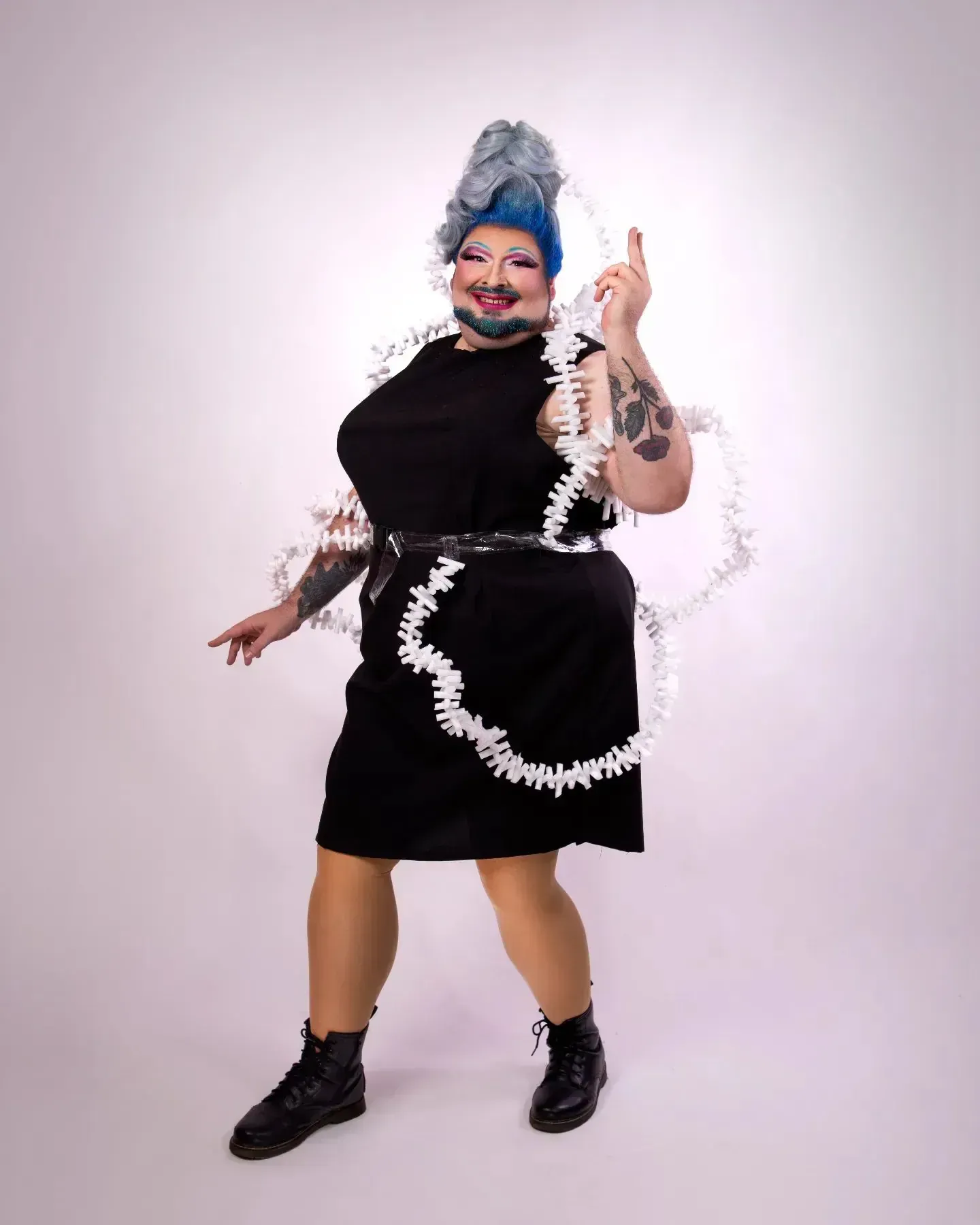 Drag performer in black dress with white trim, blue wig, raising finger, black boots.