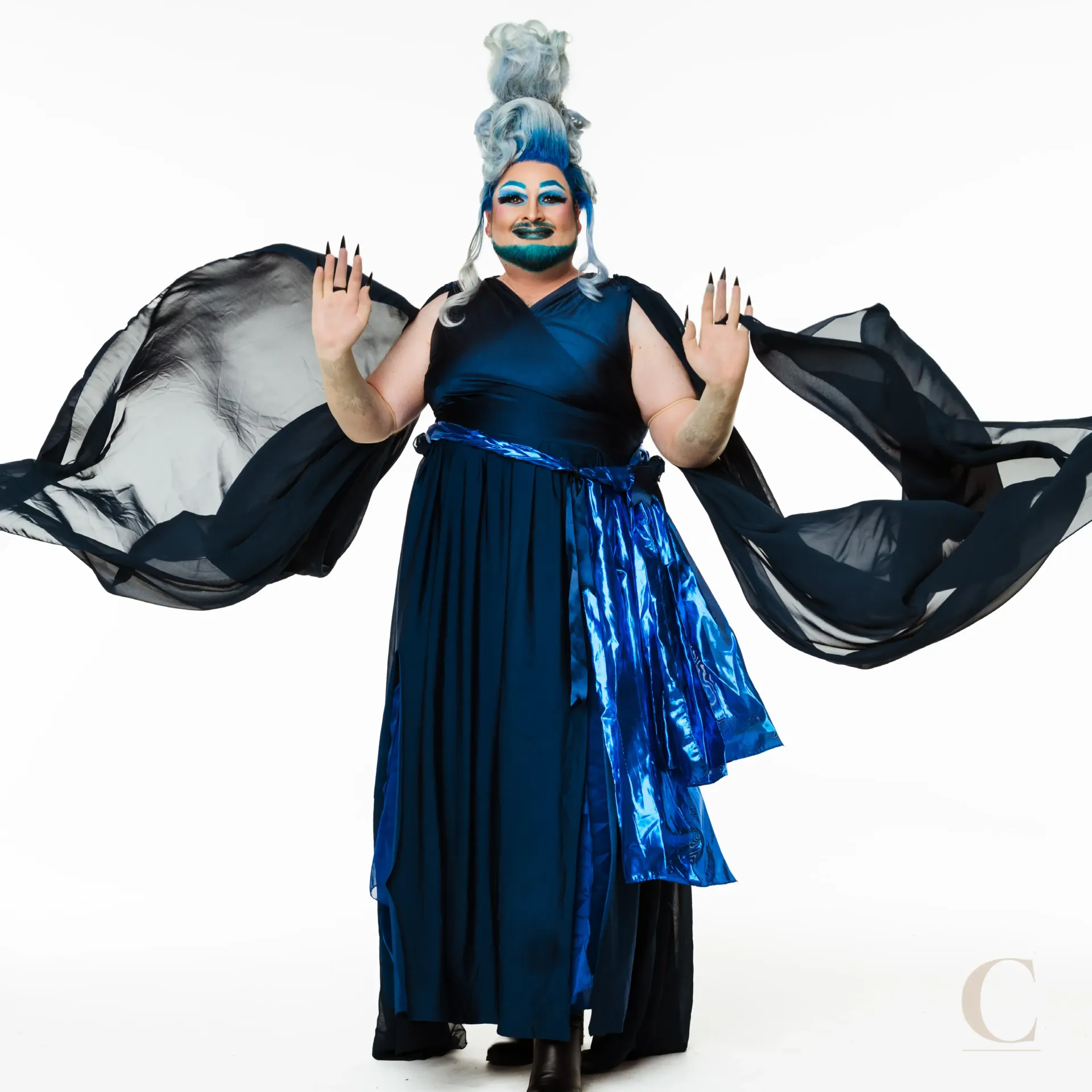 Person in a dark blue drag costume, arms raised, against a white backdrop.