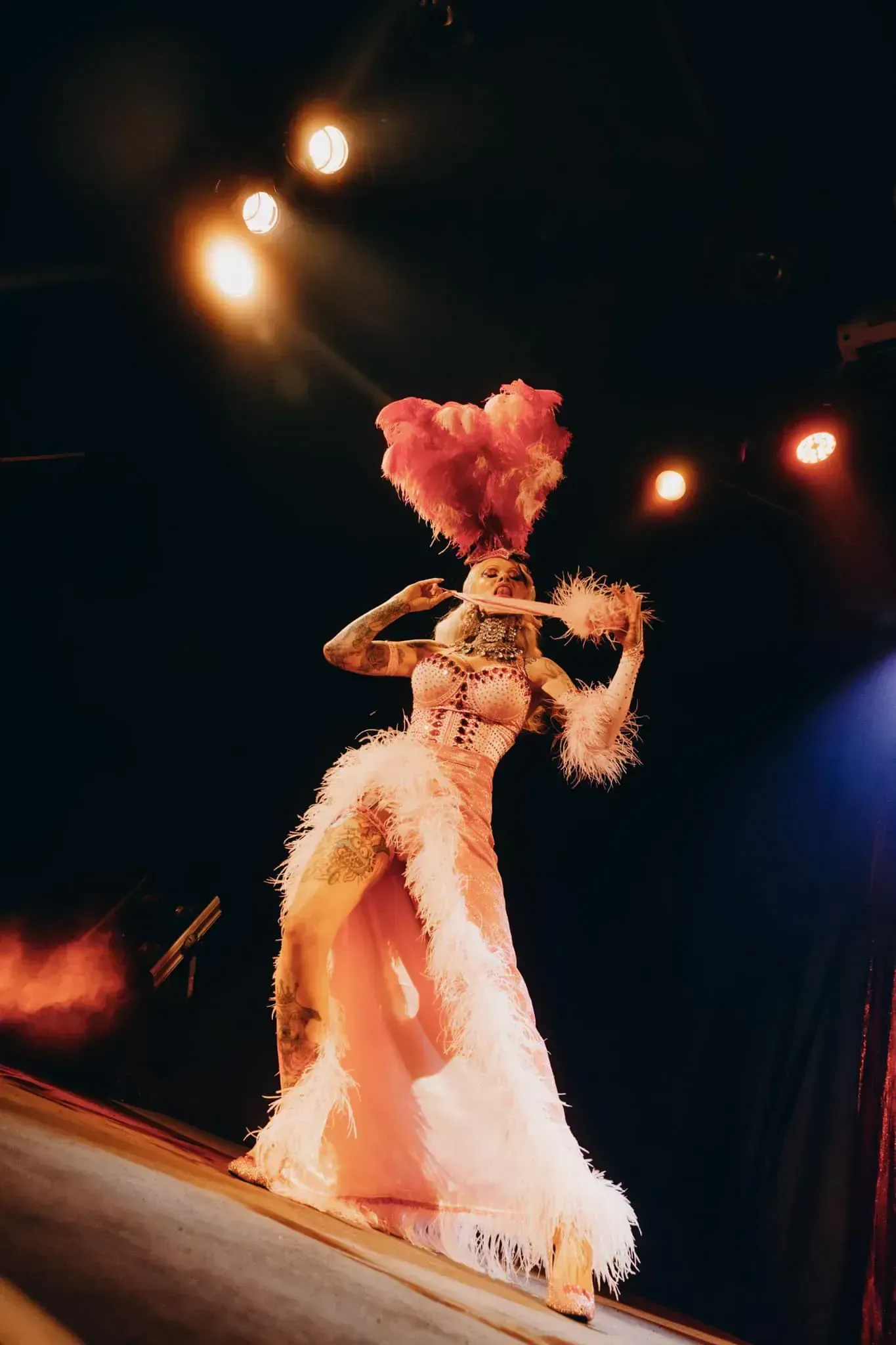Burlesque performer in light pink dress, feather headpiece stripping on stage.