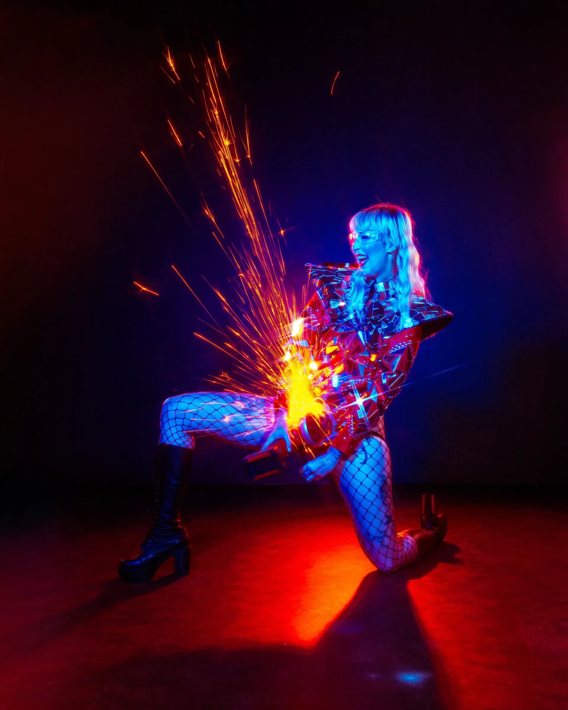 Fire dancer kneeling under neon stage lights, performing a spark shower act with angle grinder.