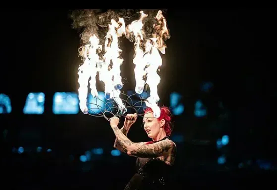 Fire dancer with red hair, holding fire fan. Performs in a dimly lit setting.