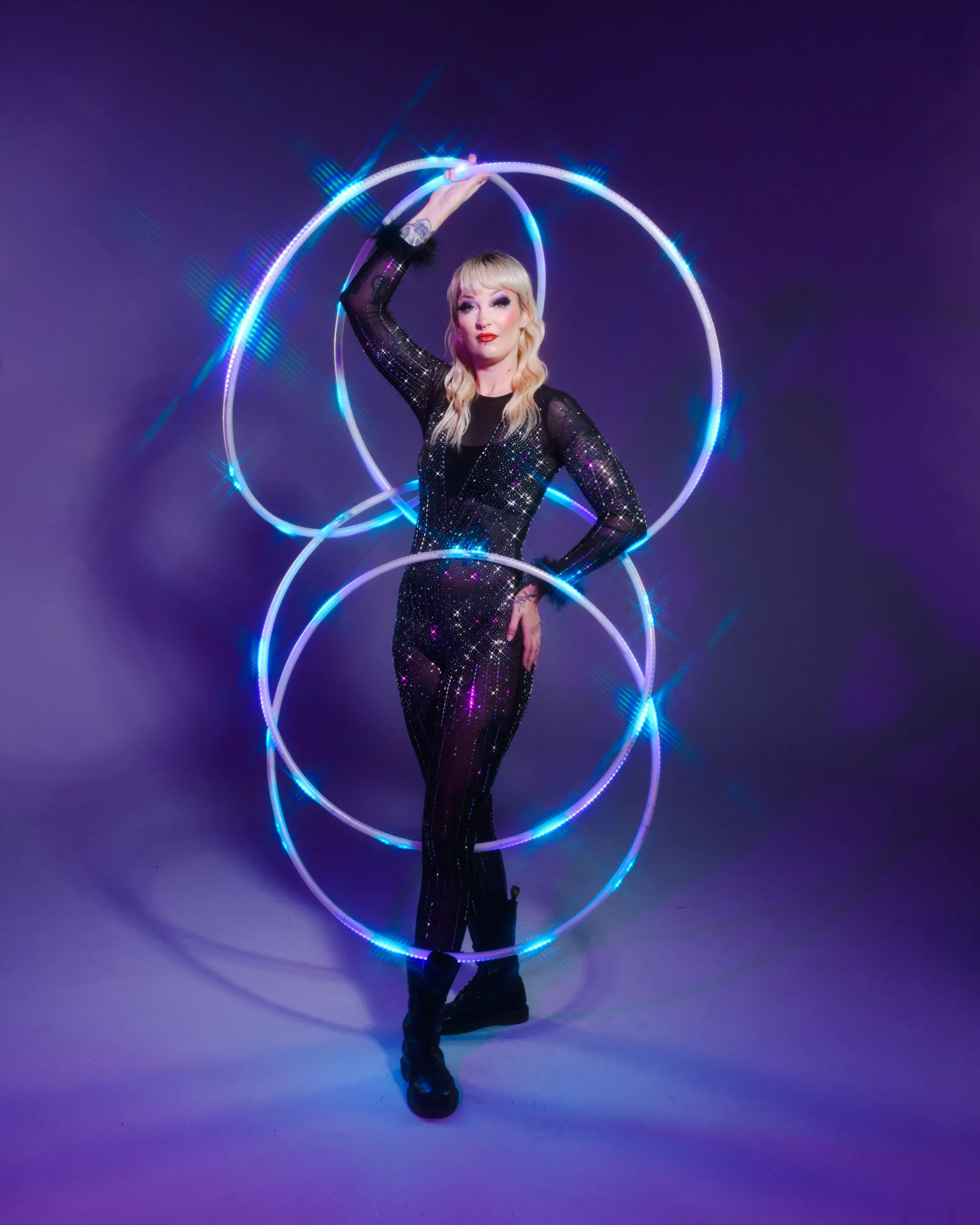 Circus artist woman holding four LED hula hoop, wearing a black bodysuit with a purple background.