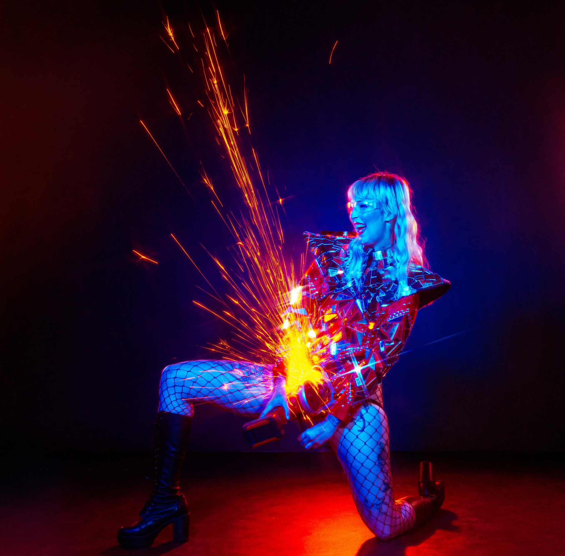 Fire dancer in silver metallic costume kneeling on grown lighting up angle grinder.