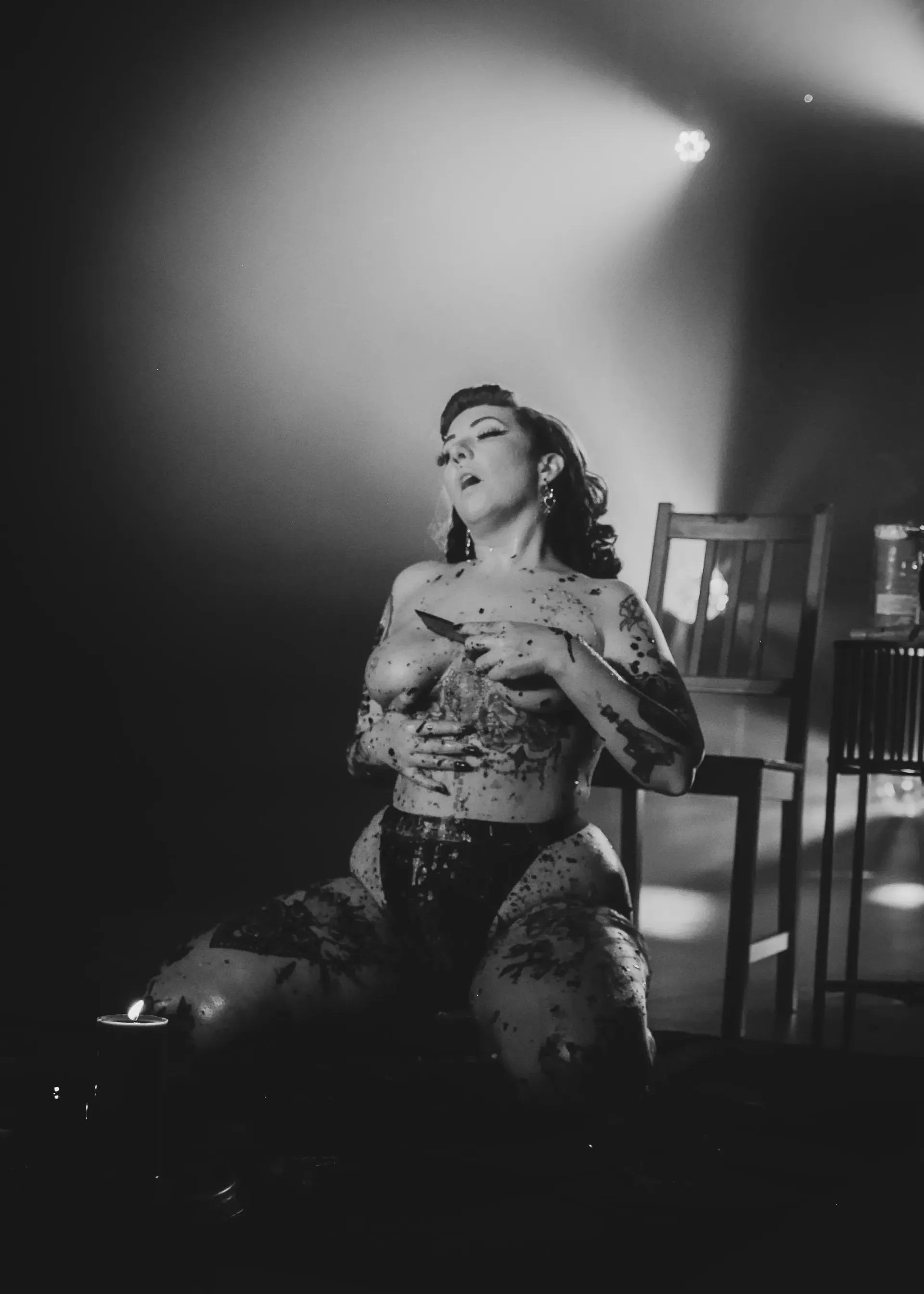 Burlesque dancer covered in dark wax, on her knees on stage, holding chest. Spotlight above, chair in background.