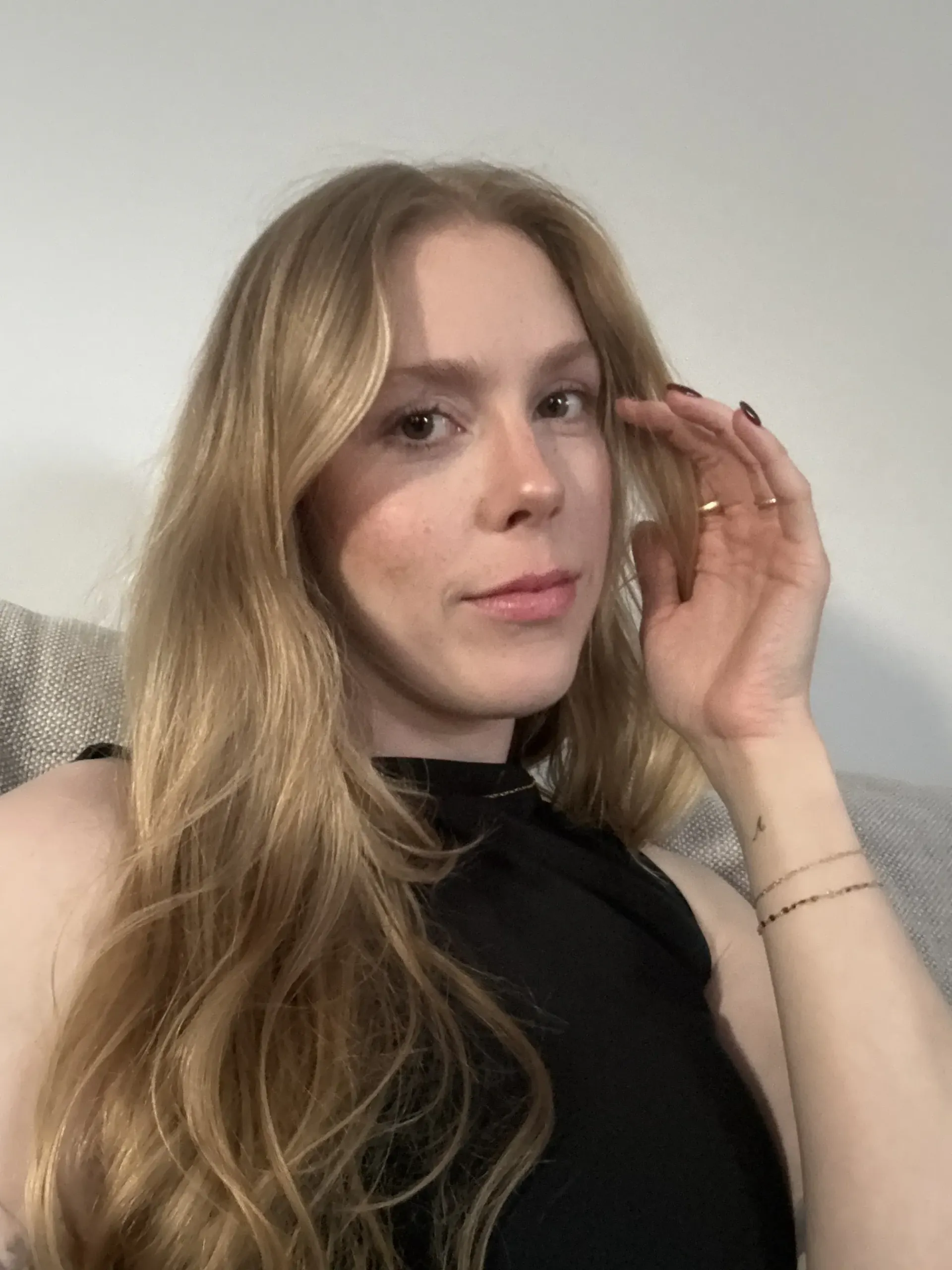 Woman with long blonde hair, touching her hair, looking at the camera. Wearing black top.