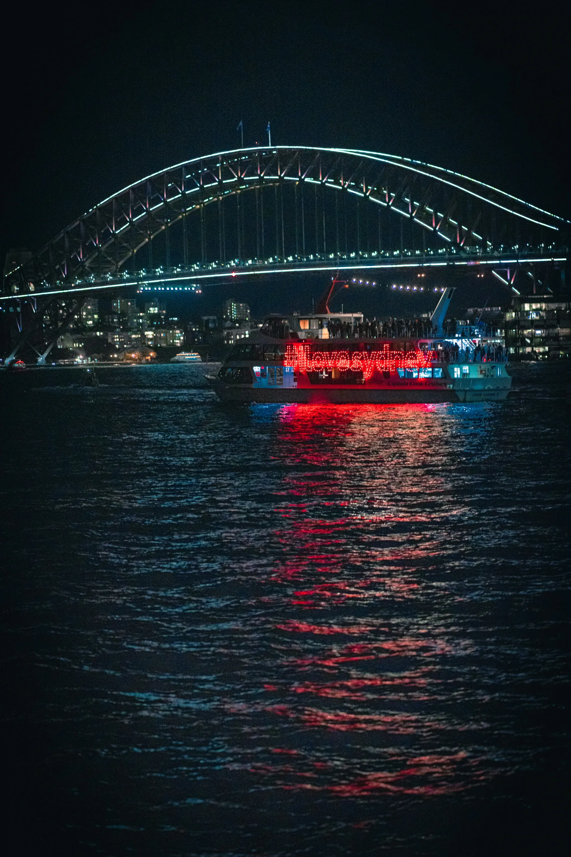 Sydney Harbour Bridge at night, lit with white lights. Red-lit boat on water.