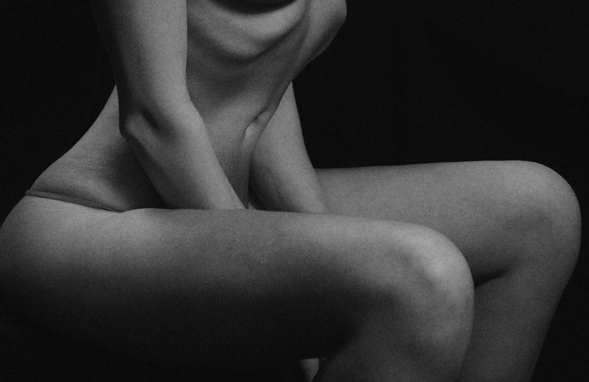 Monochrome close-up of a seated nude torso and legs, hands resting near the lap.