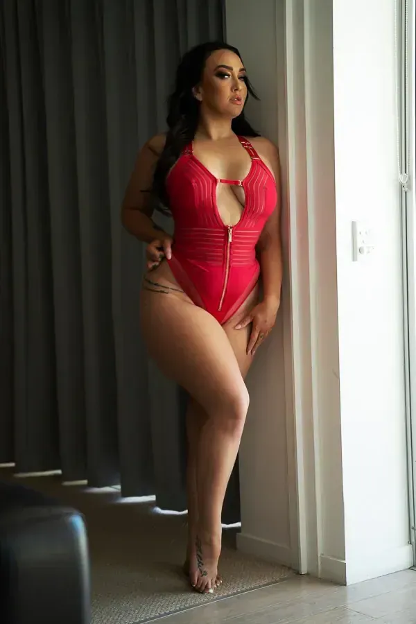 Lingerie model in red lingerie, leaning against a window frame, looking away.