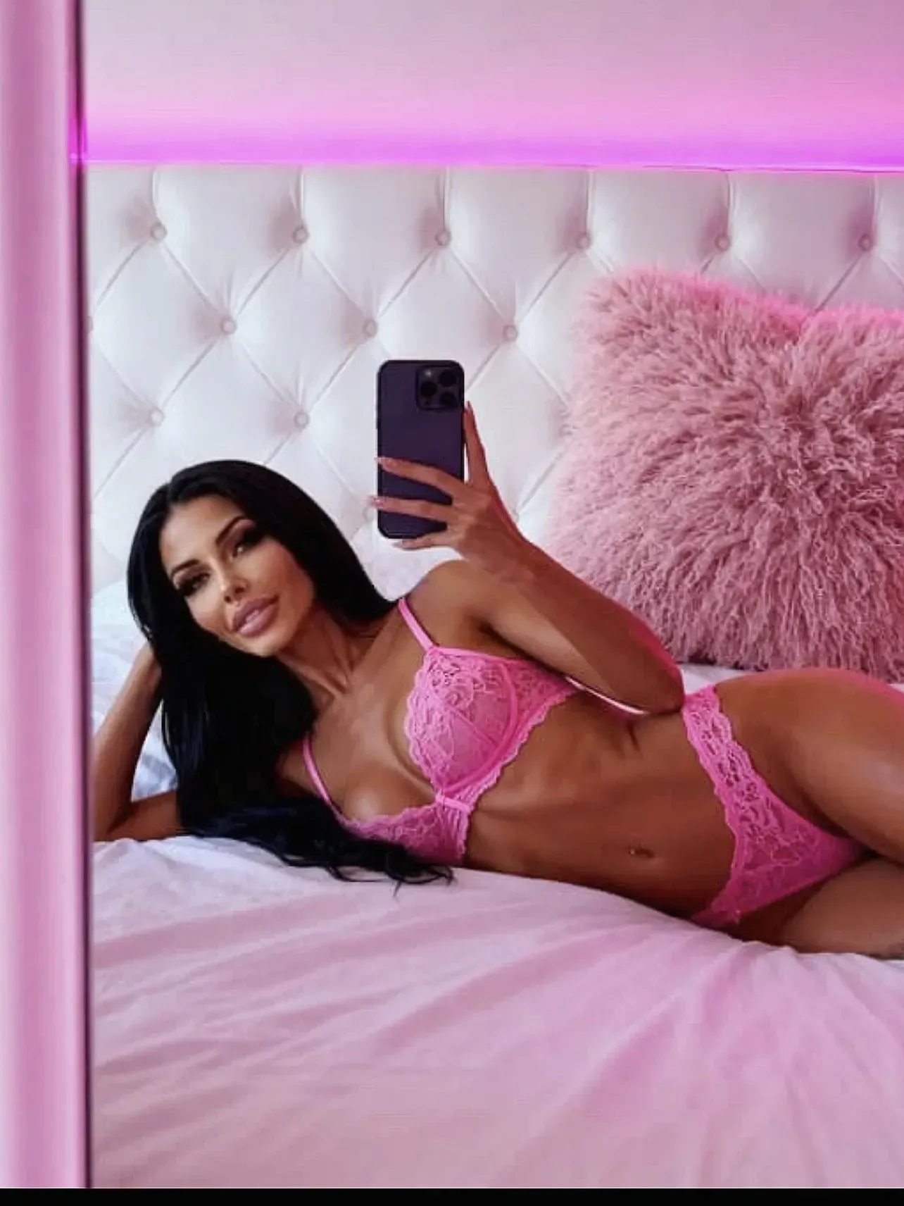Lingerie model in pink lingerie taking a mirror selfie in a pink-lit bedroom, lying on a bed with a pink headboard and pillow.