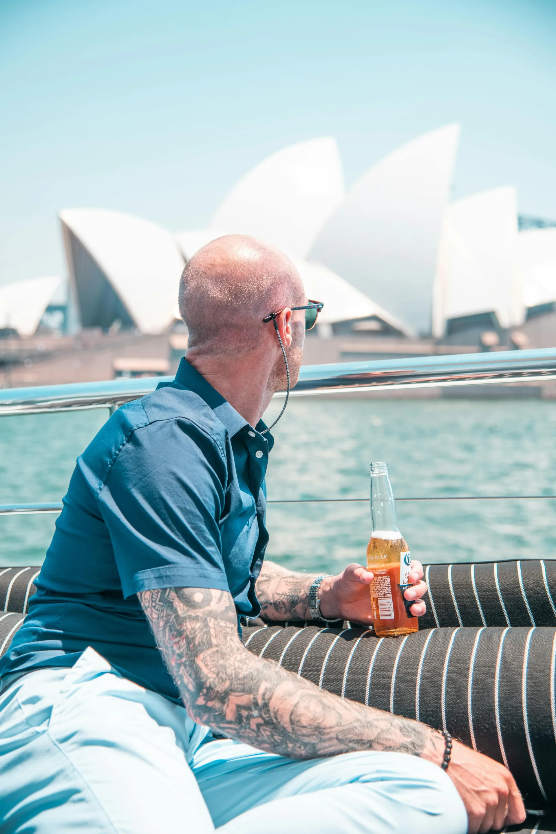 man in blue shirt holidng beer sitting and viewing Sydney Opera House.