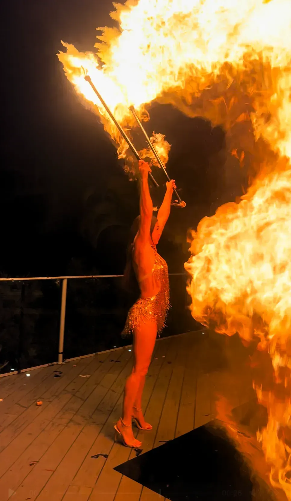 Fire dancer holding two flaming torches overhead creating a huge plume of fire erupts on outdoor deck at night.