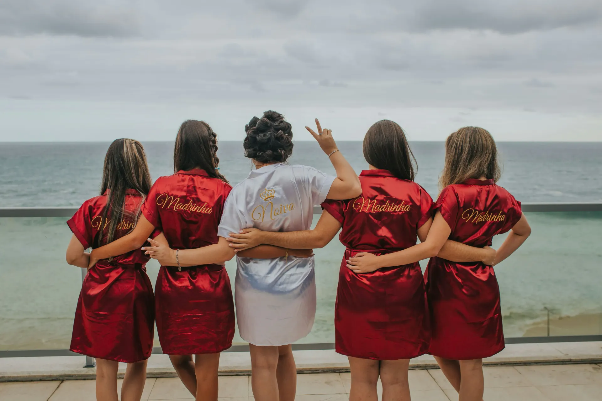 Four bridesmaids and one bride-to-be in red and white robes facing the ocean.