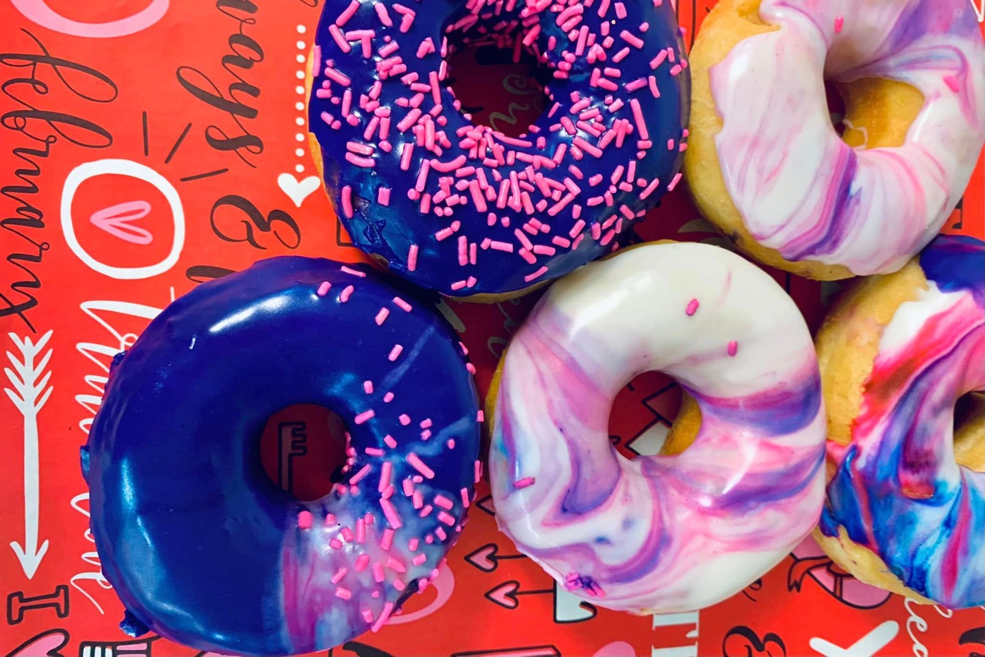 Close-up of six colorful glazed donuts on a red Valentine's Day-themed background.