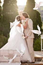 Two brides in white gowns share a kiss outdoors during a wedding ceremony.