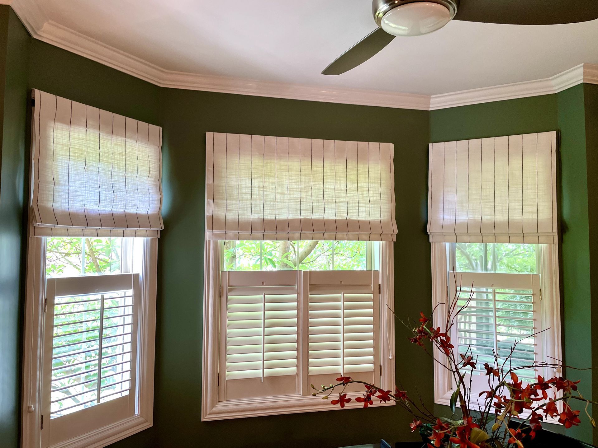 Three windows with white shutters and linen Roman shades on a green wall. A ceiling fan is above.
