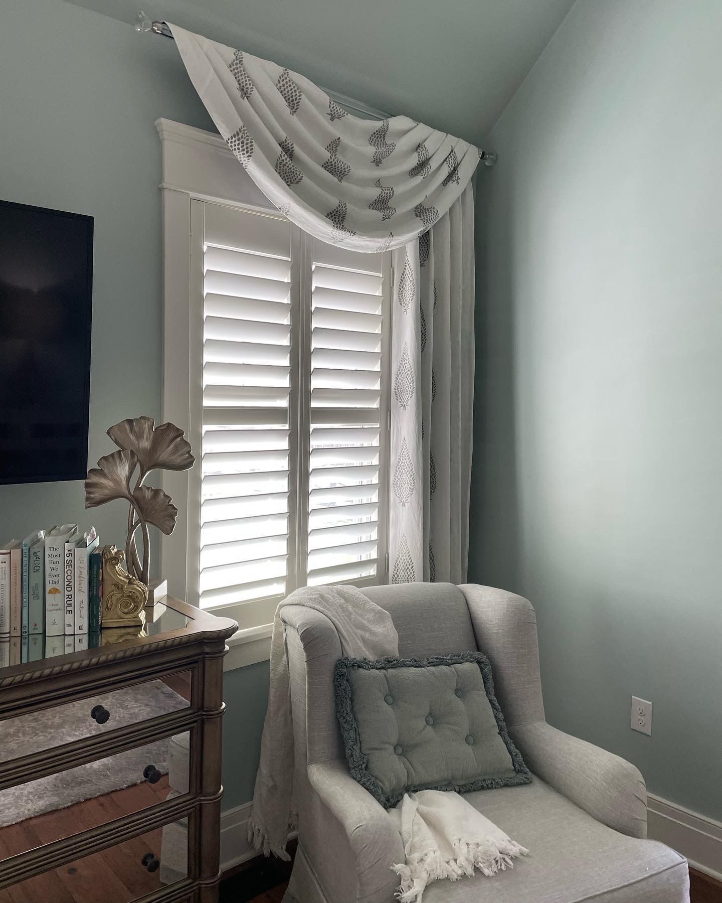 Cozy reading nook with white shutters, chair, patterned curtain, and a small table with a vase.