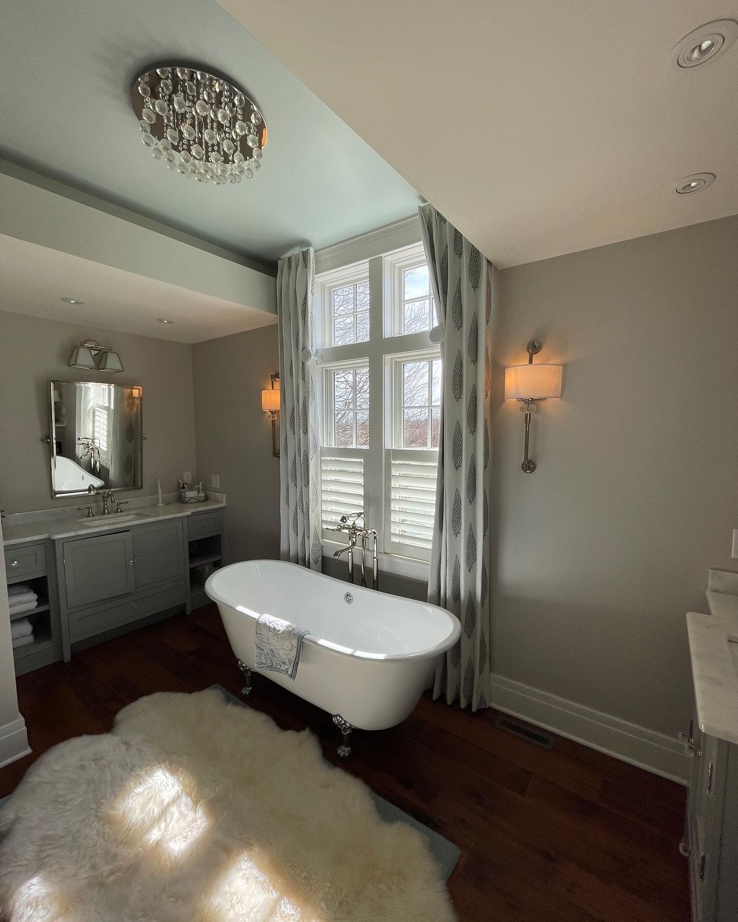 Bathroom with freestanding tub, vanity, chandelier, and window with curtains.