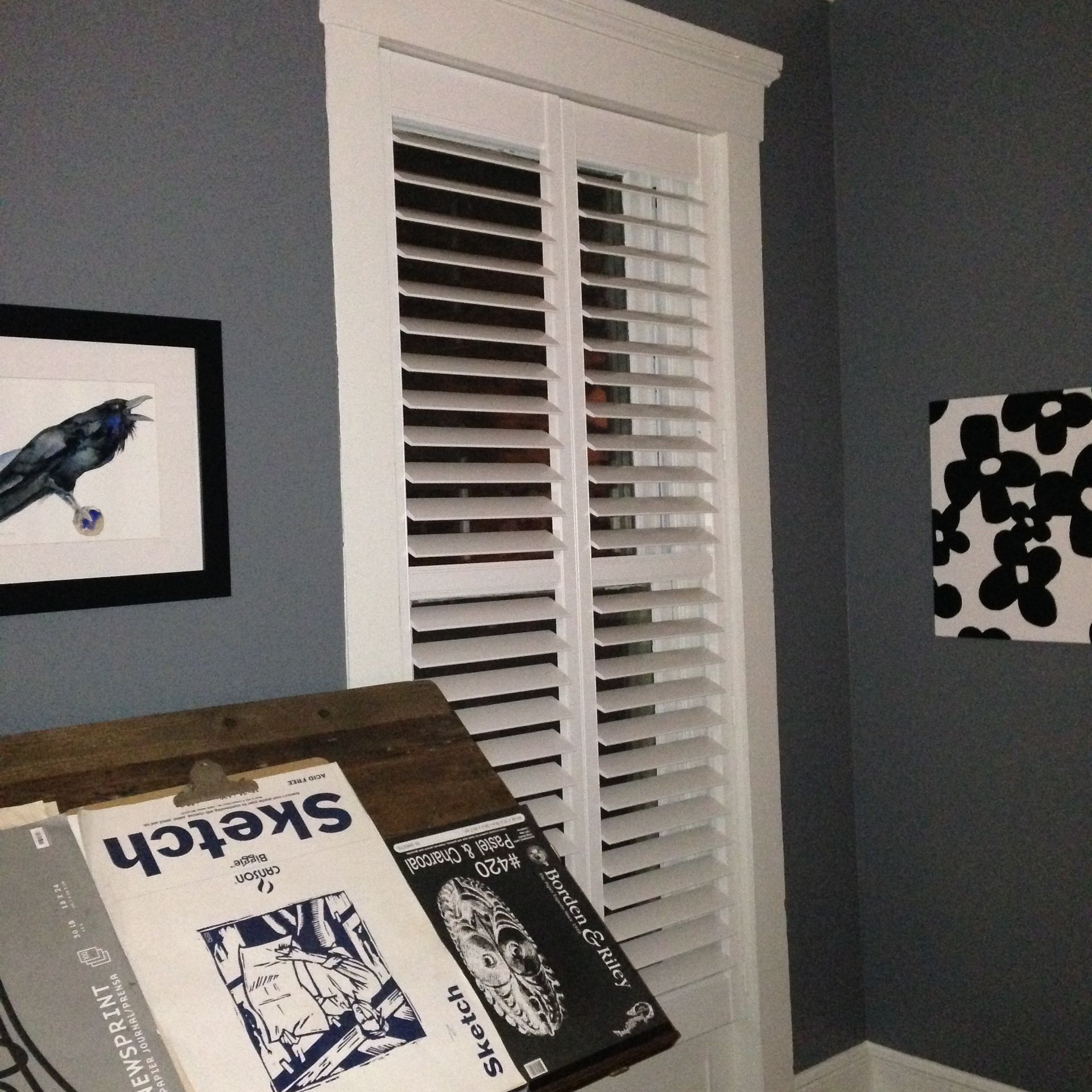 White shutters on a window in a gray-walled room; art and sketchbooks are also present.