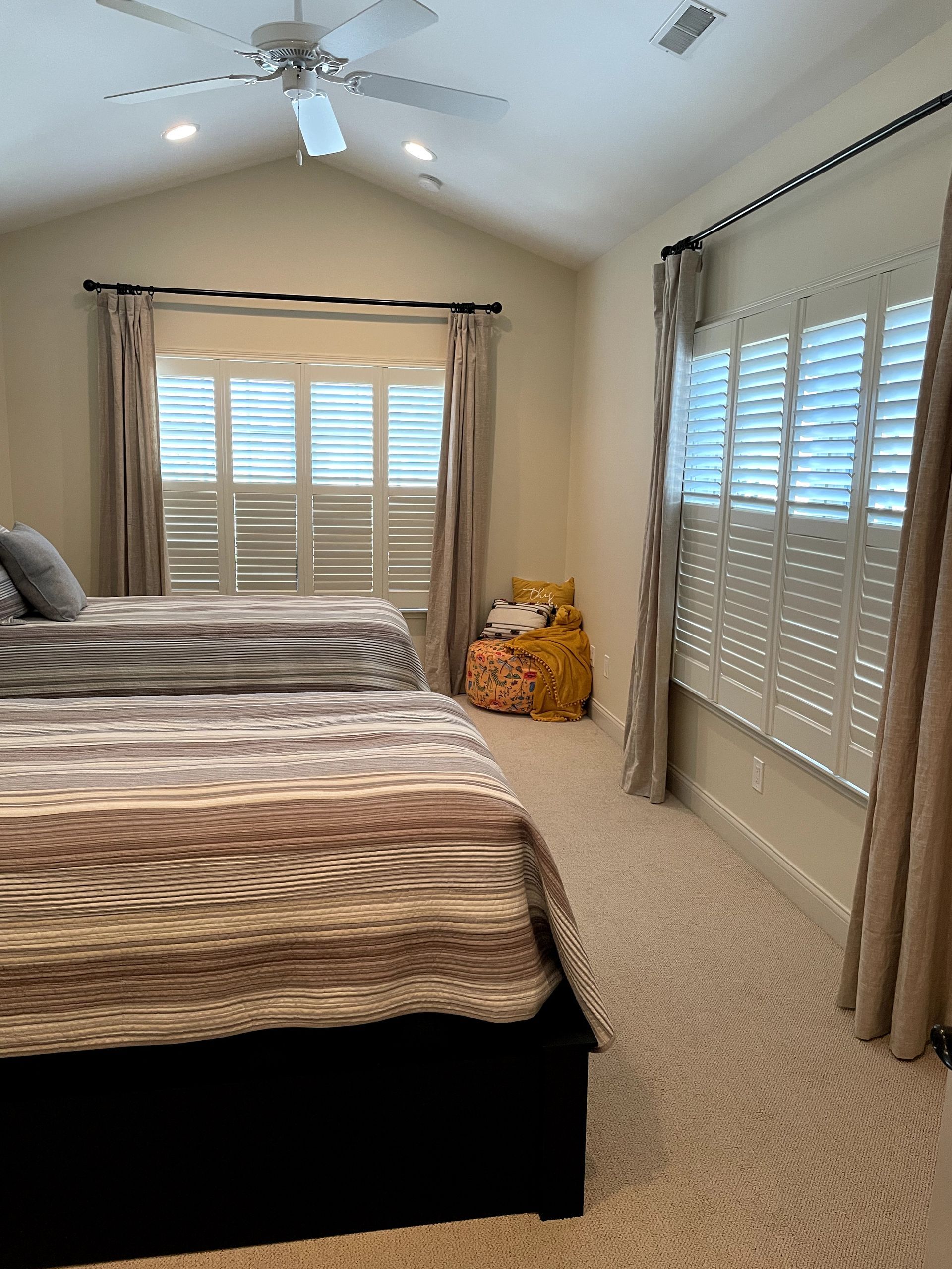 Two beds in a light-filled room with window shutters, beige carpet, and neutral-toned bedding.