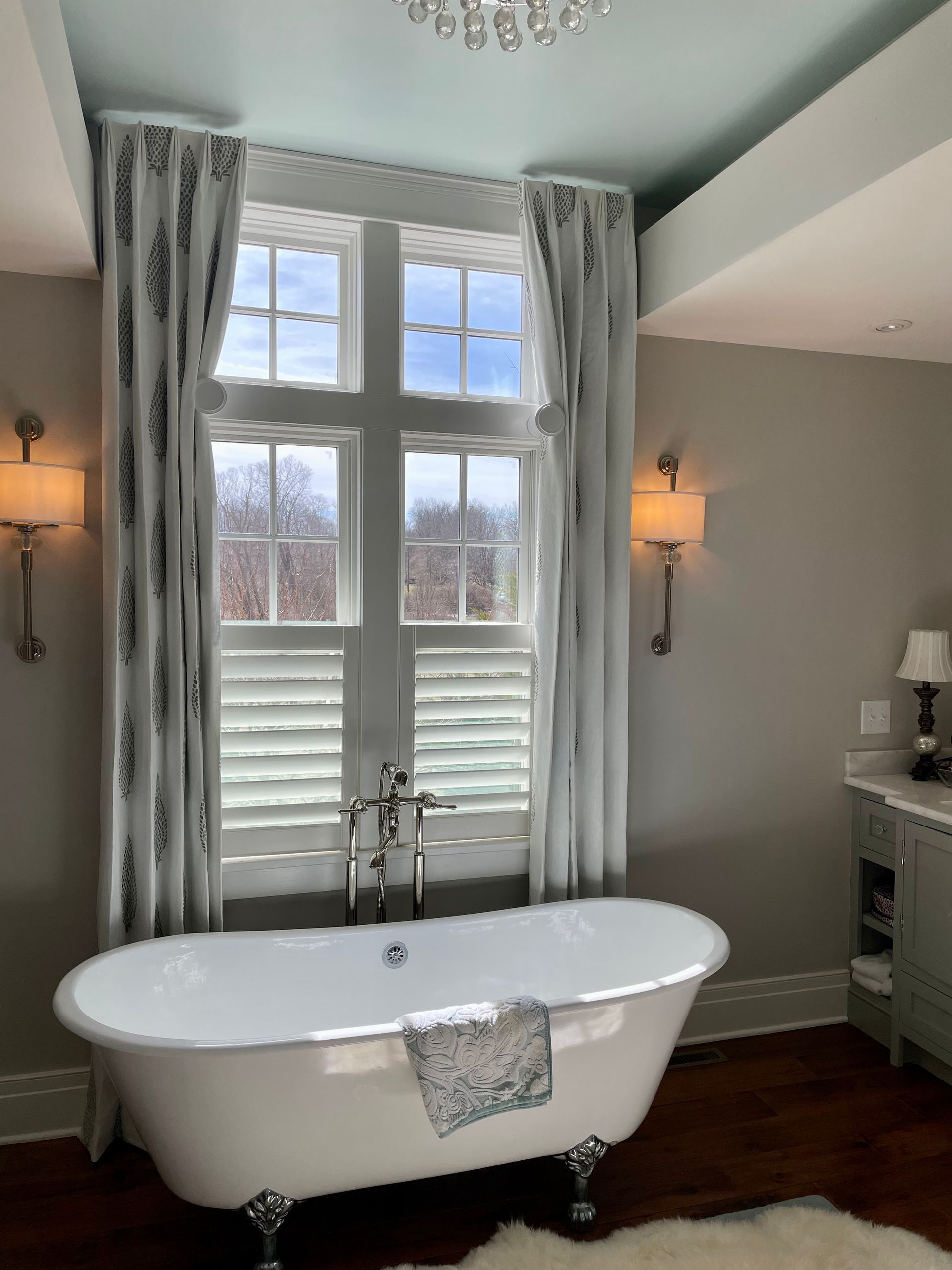 Clawfoot bathtub in a bathroom with tall window, curtains, wall sconces, and light blue ceiling.
