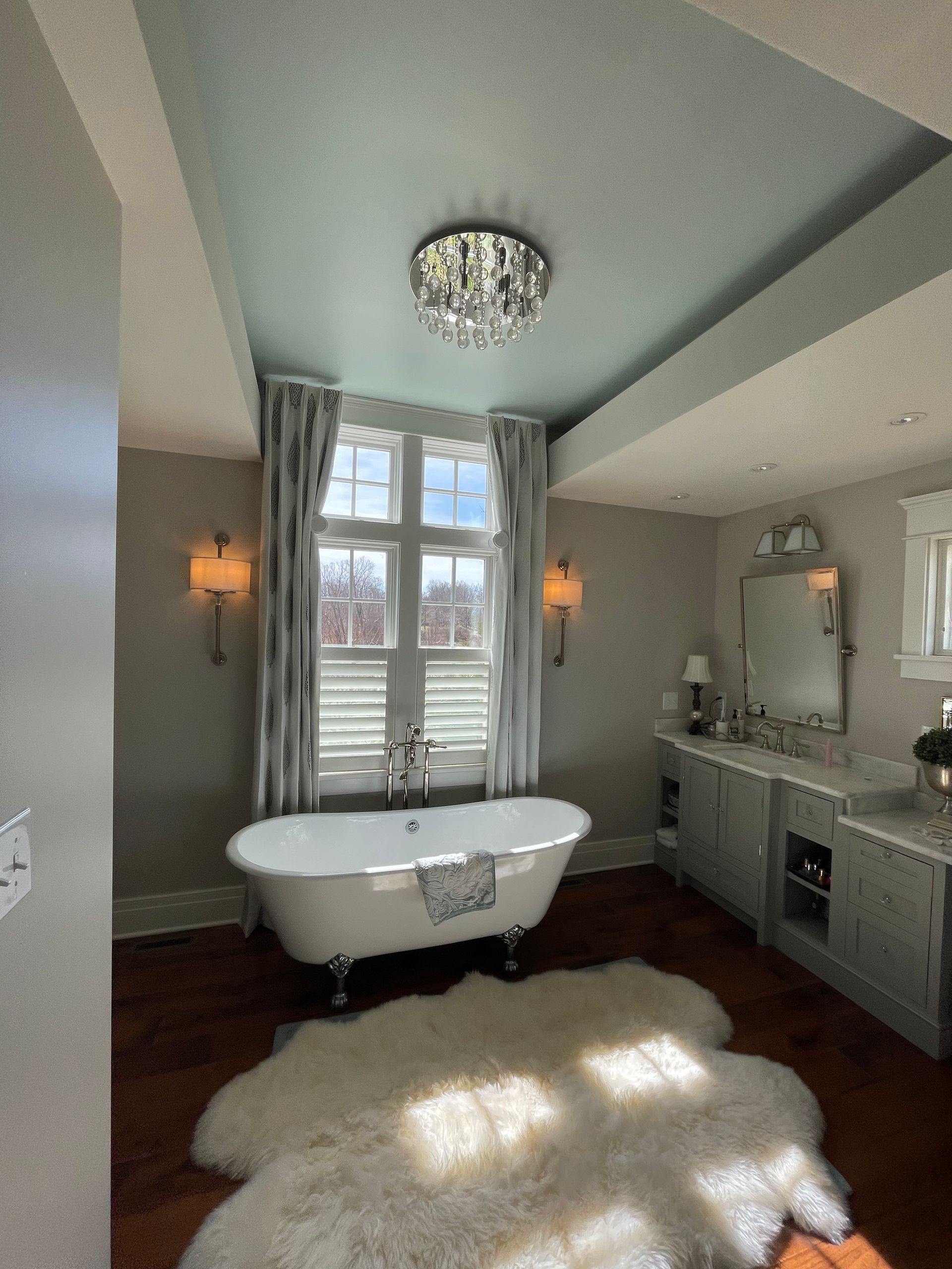 Bathroom with clawfoot tub, light blue ceiling, chandelier, window with curtains, and a fluffy rug.